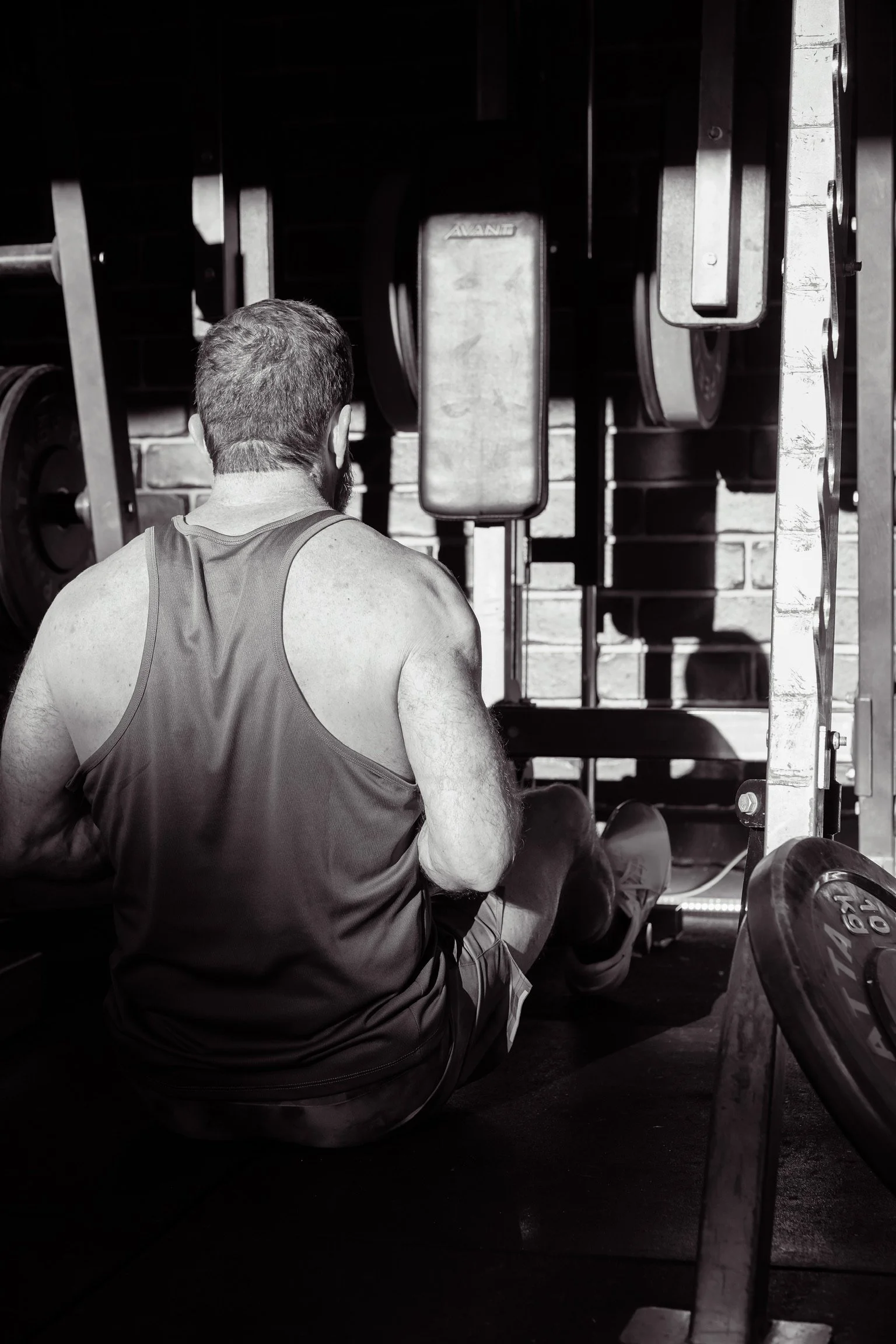 A man with short hair and a beard, wearing a sleeveless athletic shirt, is sitting on the floor of a gym in front of a large weightlifting mirror, preparing for a workout. There are various weights and gym equipment visible around him.