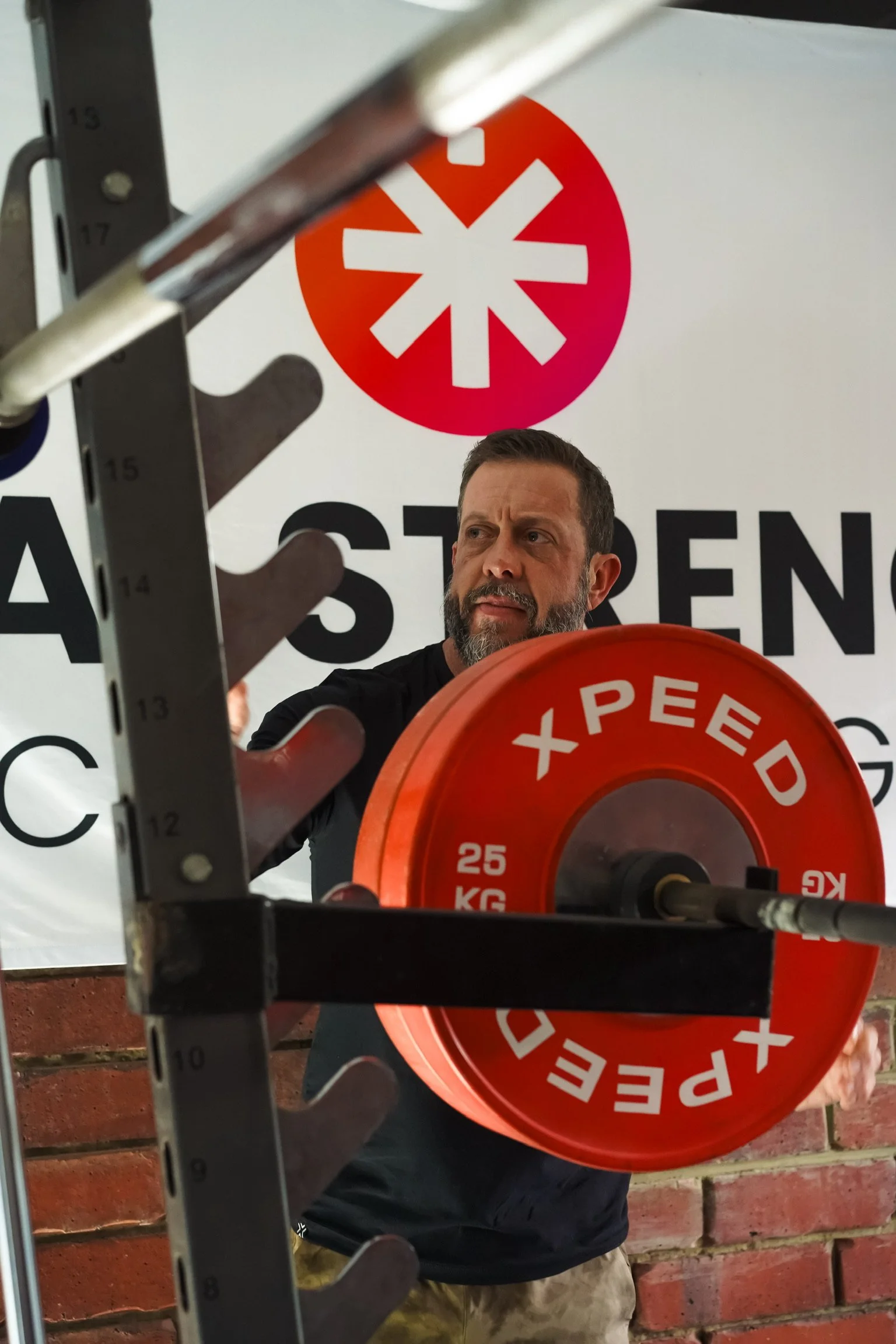 A man with a beard lifts a red weight plate in front of a white background with red and black signage, partially visible, with a measuring scale and a hand in the foreground.