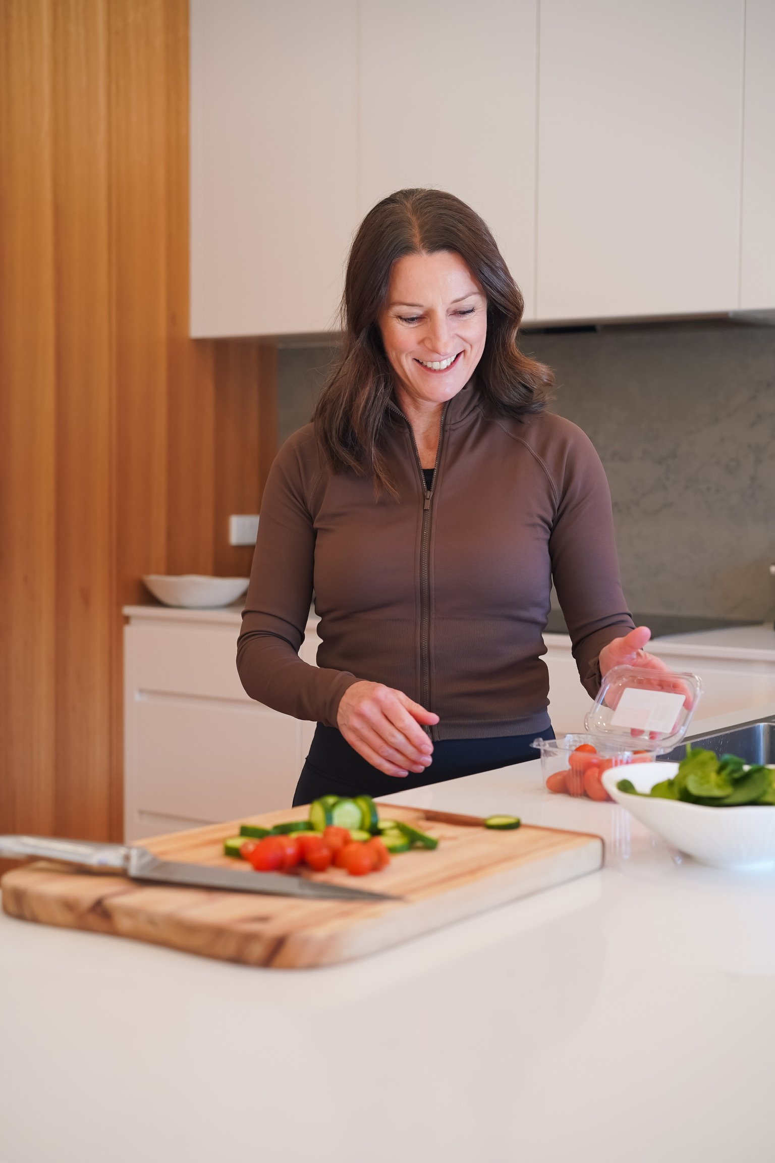 A woman with brown hair smiling while preparing vegetables on a kitchen counter. There are sliced cucumbers, cherry tomatoes, and carrots, along with bowls of greens and whole carrots. The kitchen features wooden and white cabinets.