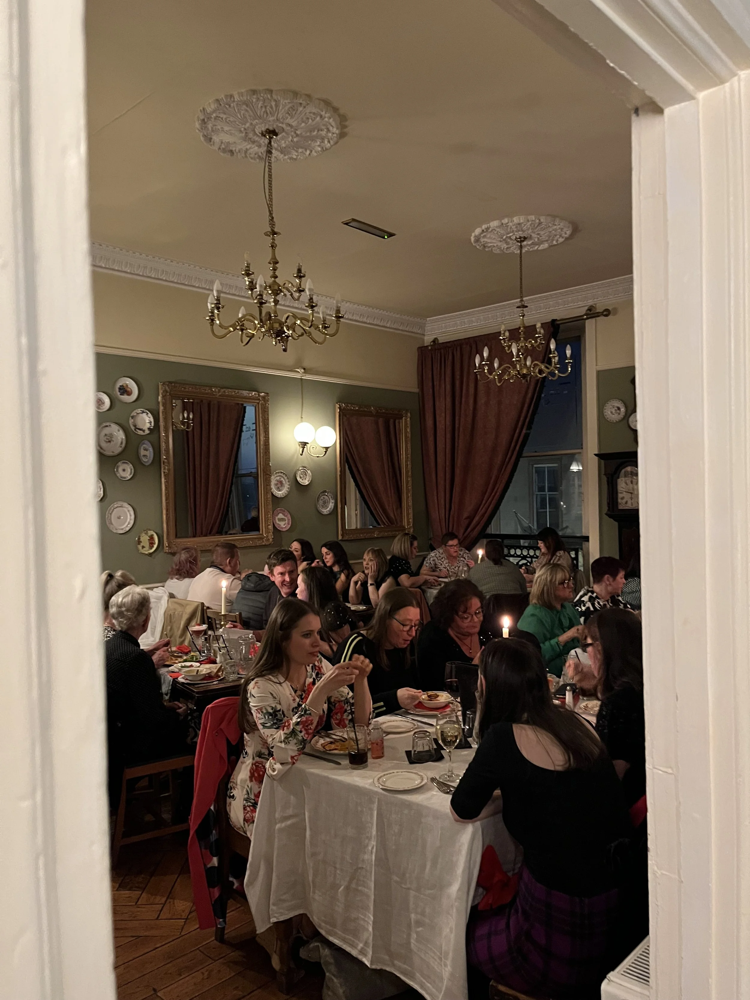 A dining room filled with people enjoying a meal together. The room features elegant chandeliers, framed mirrors, decorative plates on the wall, and large red curtains. The table is set with dishes, glasses, and lit candles.