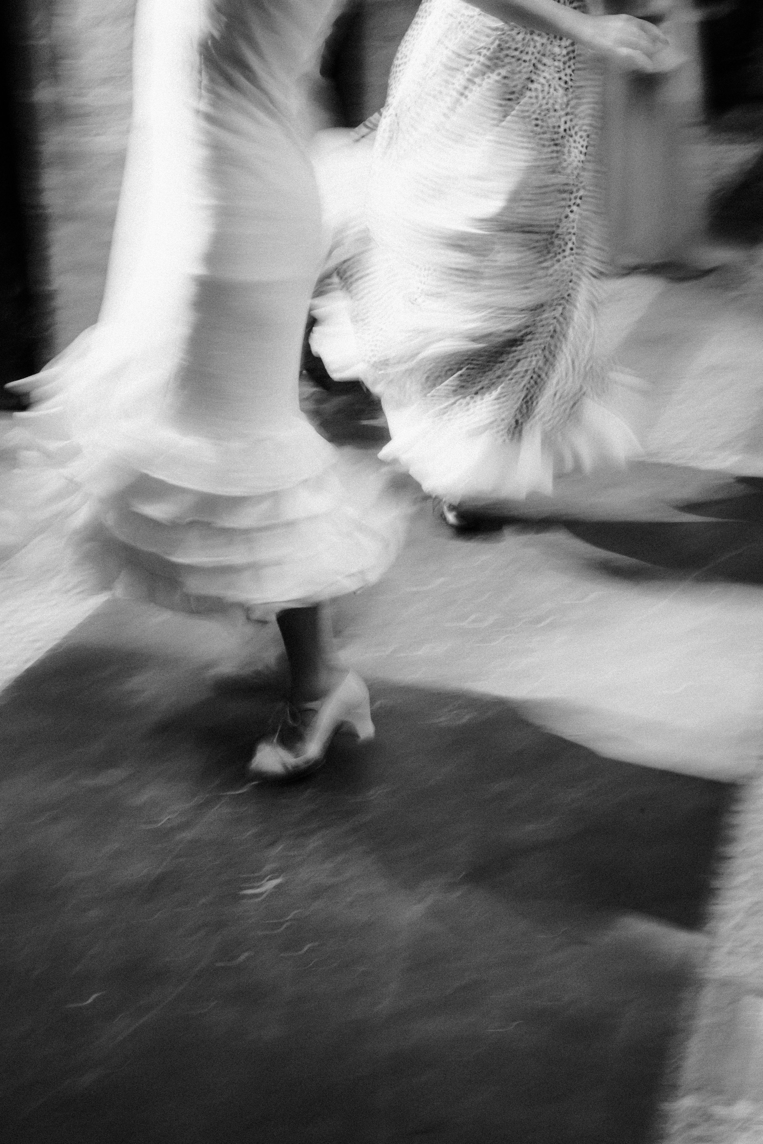 Blurred black and white image of two people dancing, showing movement and flow of dresses and shoes.