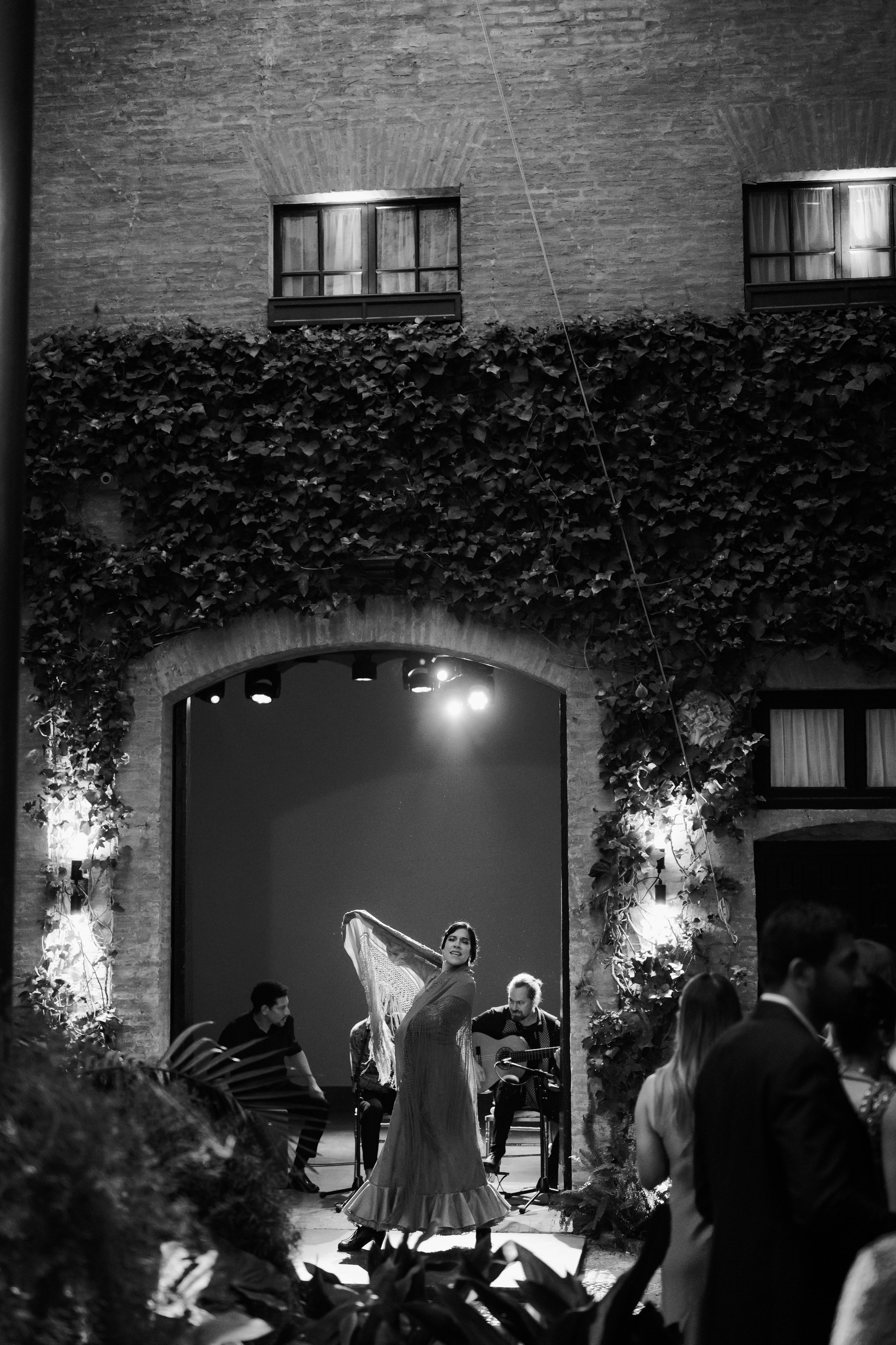 Black and white photo of a flamenco dancer performing in a courtyard, accompanied by musicians playing guitar. The setting features brick walls and ivy, with an audience watching.