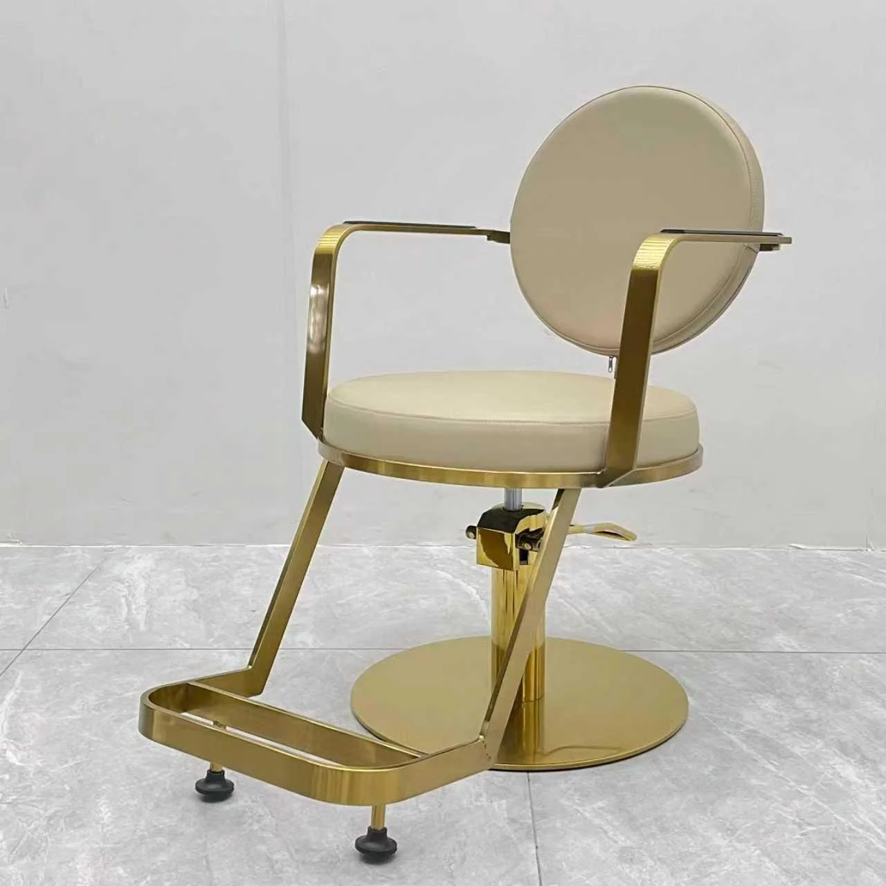 A modern salon chair with a gold frame, beige cushioned seat and backrest, and a footrest, set against a plain light-colored background.