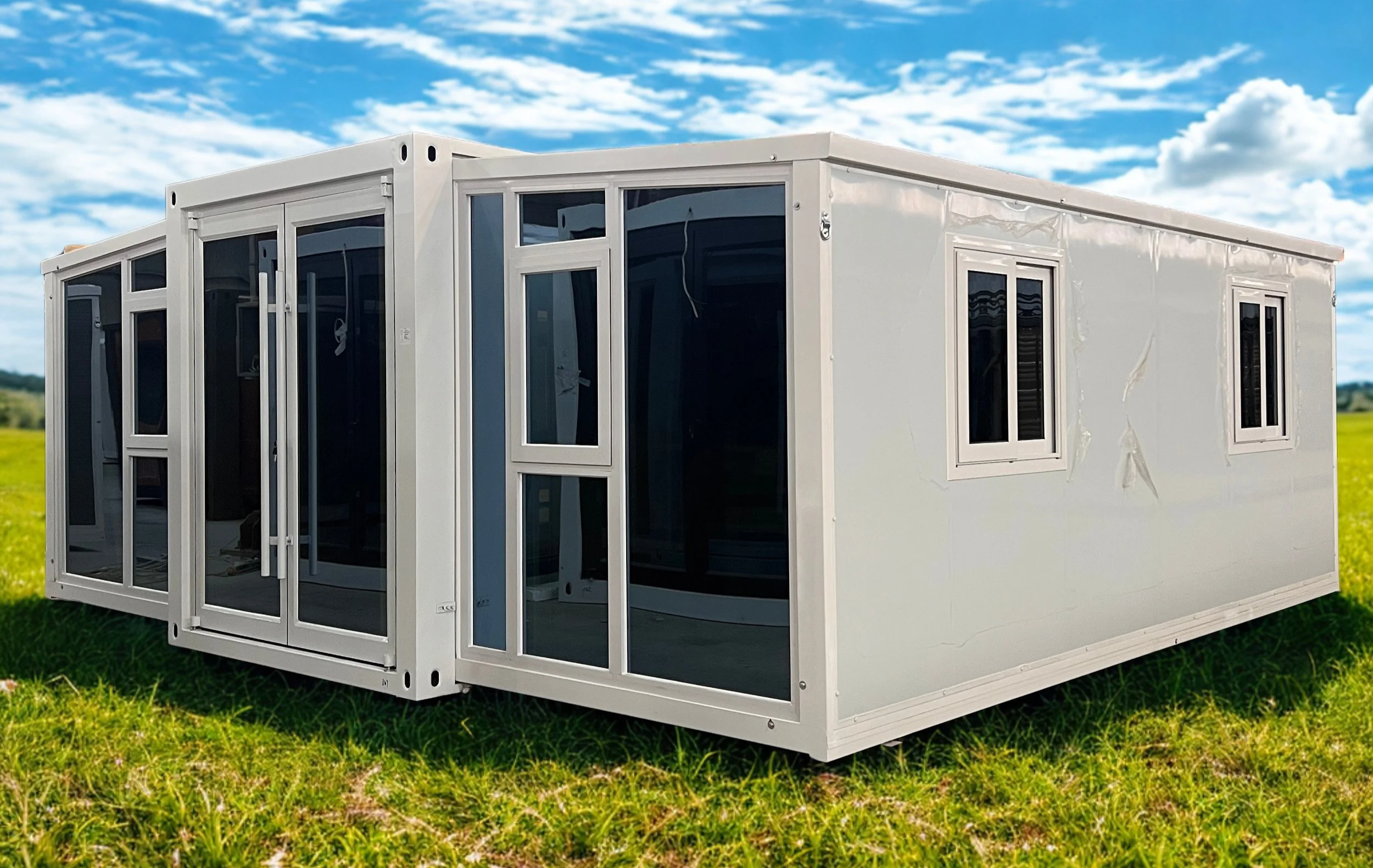 A white modular prefabricated building with large glass windows and sliding glass doors, placed on a grassy field under a blue sky with scattered clouds.