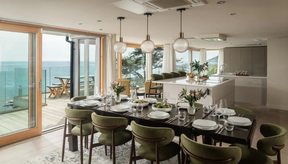 Modern open-plan dining and kitchen area with large windows overlooking the ocean, decorated with flowers, table set with white plates and glassware, and a small balcony outside.