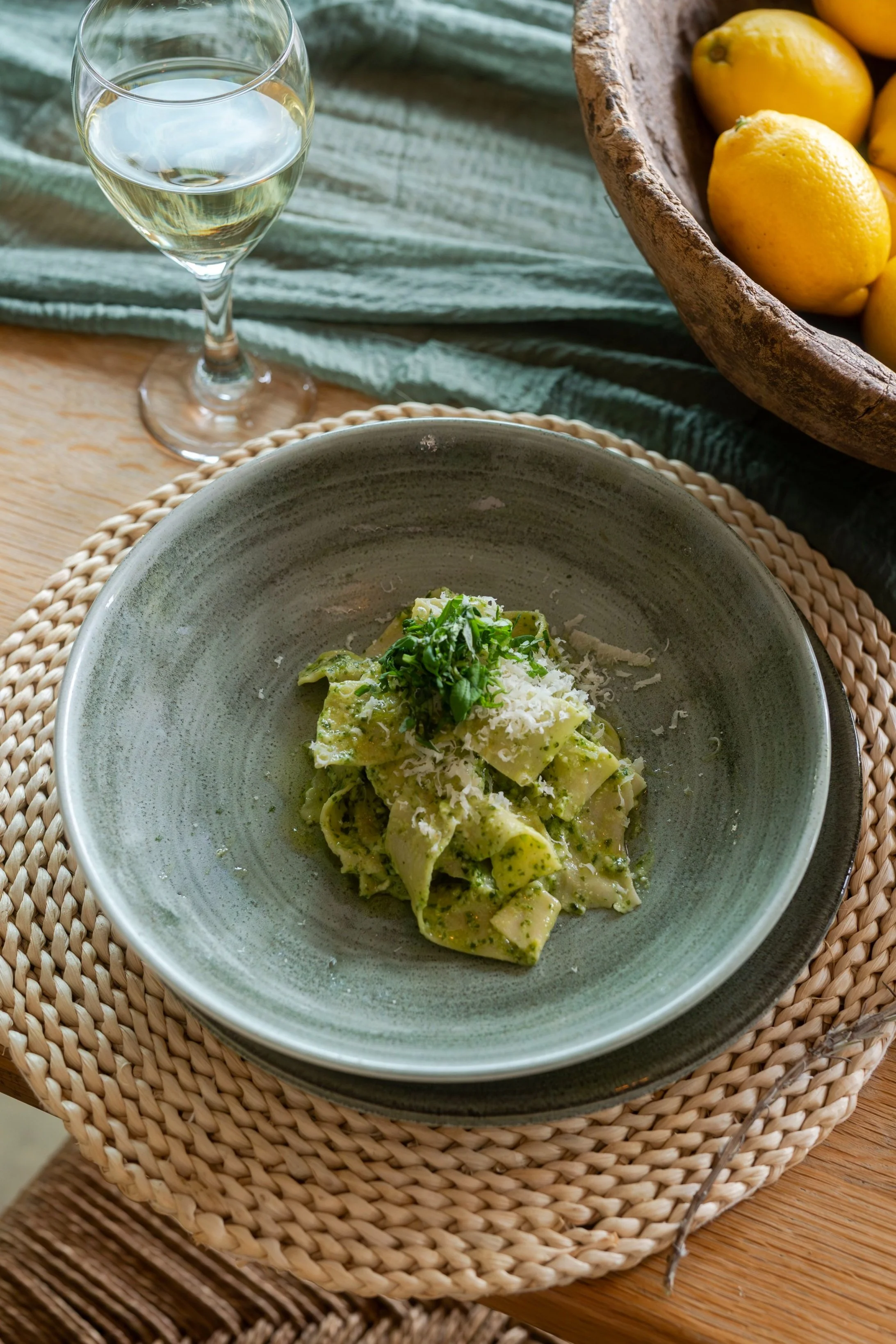 A plate of green pesto pasta topped with grated cheese and fresh herbs on a woven placemat, alongside a glass of white wine and a bowl of lemons on a rustic wooden table.