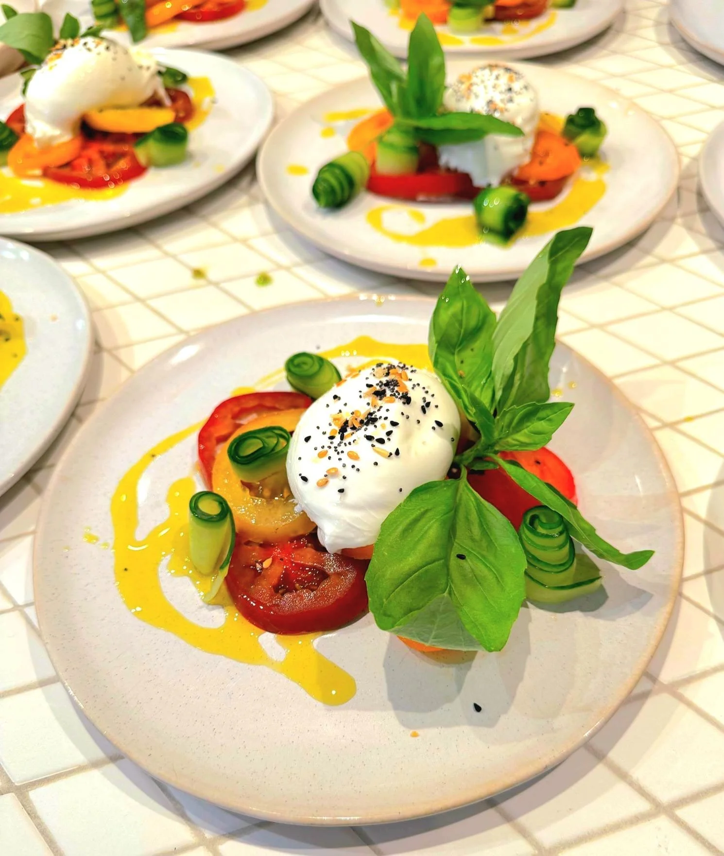 Caprese salad with sliced tomatoes, fresh basil, mozzarella cheese, drizzled with olive oil, and sprinkled with black sesame seeds, served on a white plate.