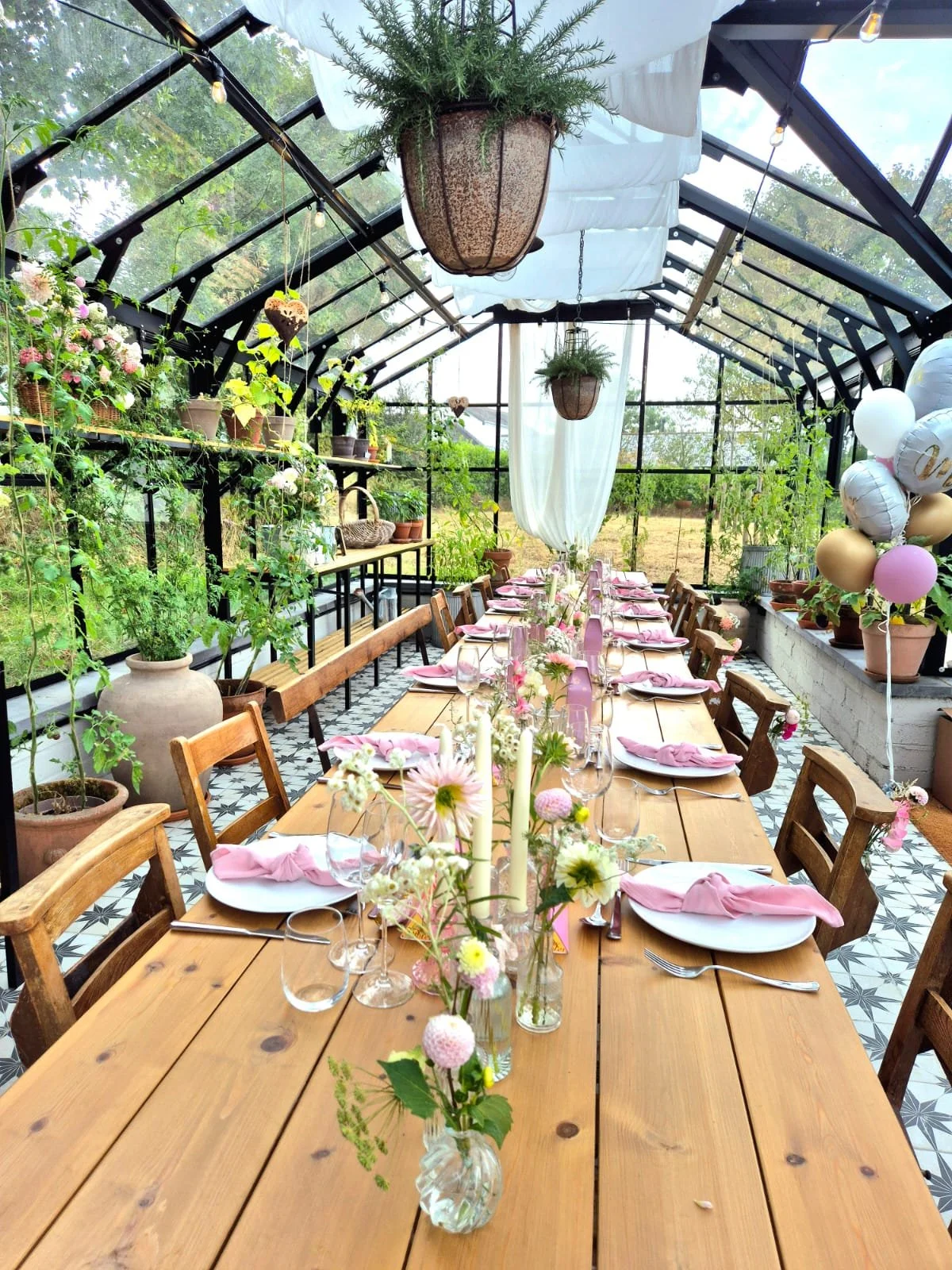 Long wooden table set for a celebration inside a glass greenhouse, decorated with pink napkins, glassware, and flowers in vases, with hanging plants and balloons in the background.