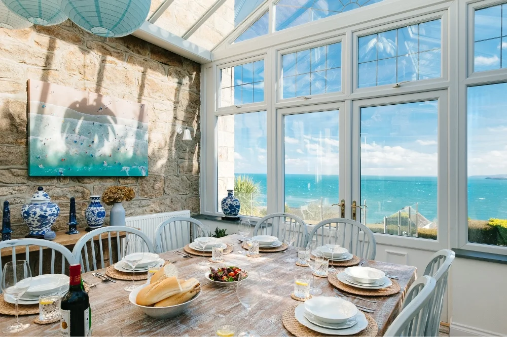 A dining room with a large wooden table set with plates, glasses, bread, and a pitcher of water. The room has large windows showing a view of the ocean, blue sky with clouds, and a coastline.