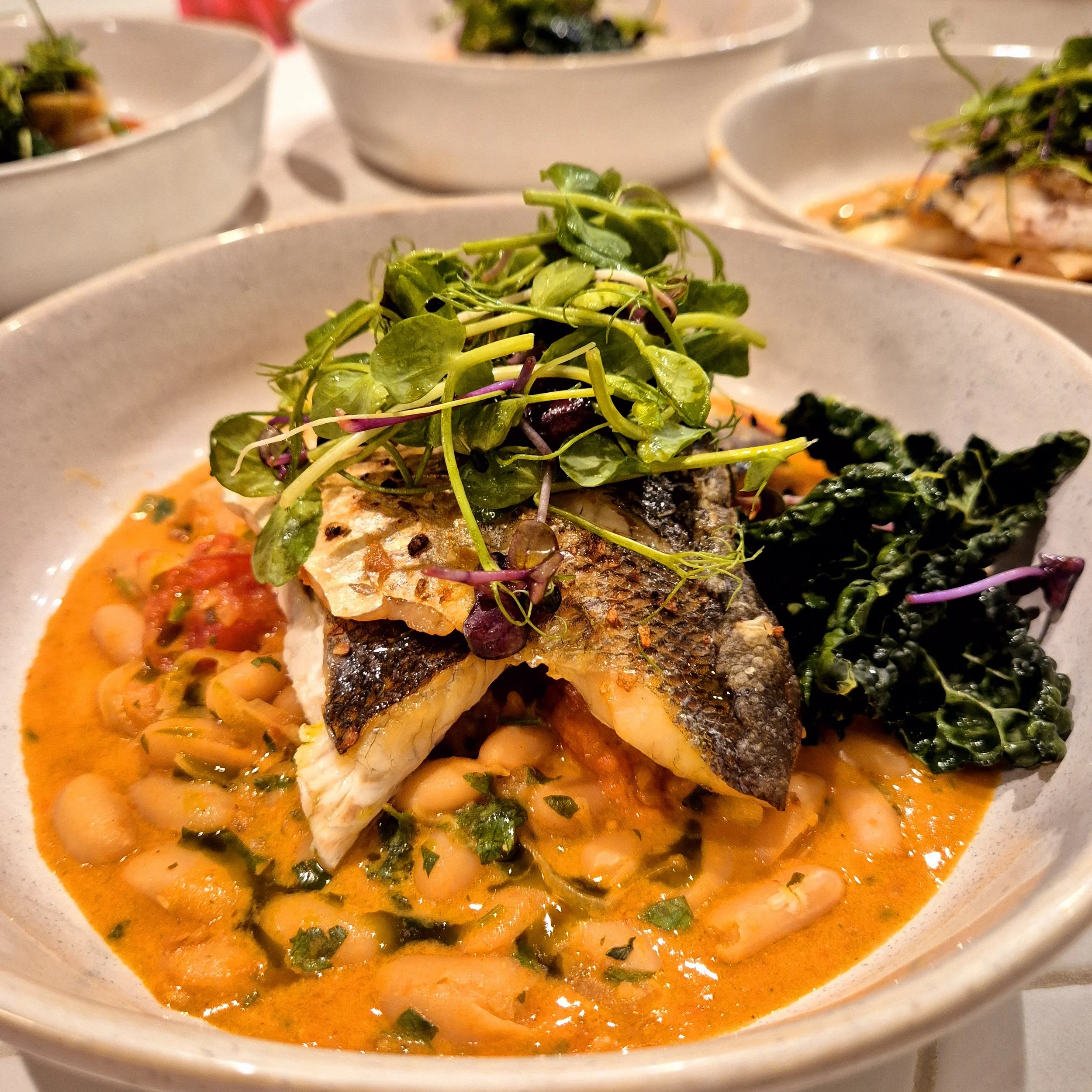 Gourmet dish with grilled fish on a bed of white beans in tomato sauce, topped with microgreens, beside sautéed greens, served in a white bowl.