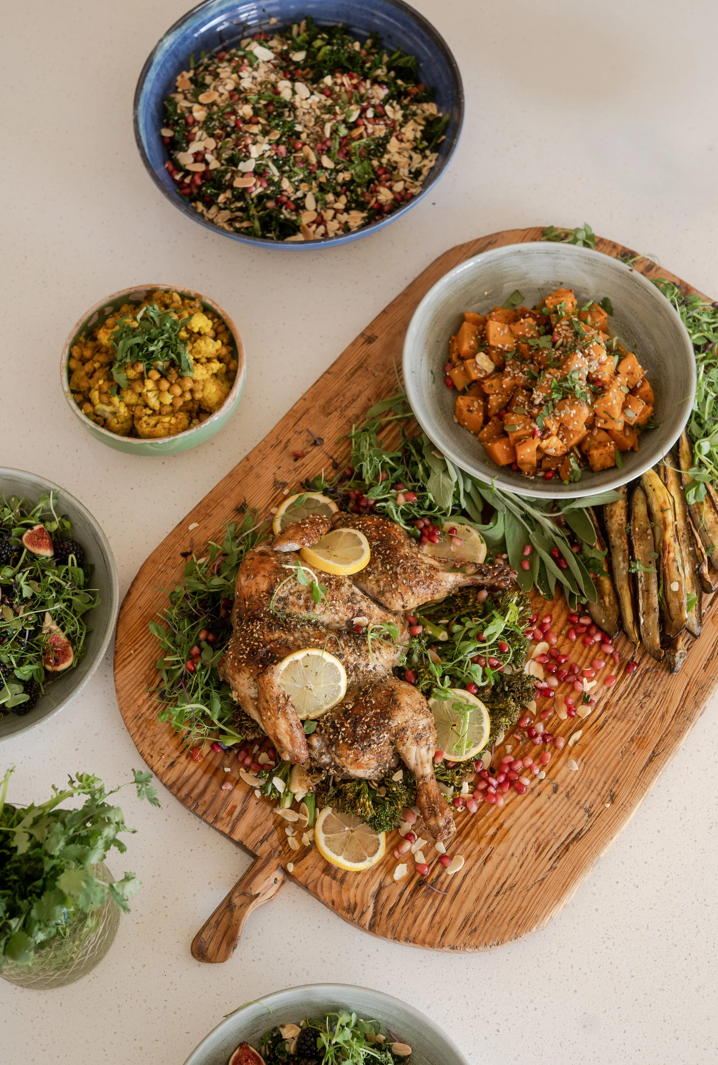 A roasted chicken garnished with lemon slices and herbs on a wooden board surrounded by bowls of salads and vegetables, including roasted sweet potatoes, mixed greens with seeds and nuts, spiced chickpeas, and grilled eggplant.