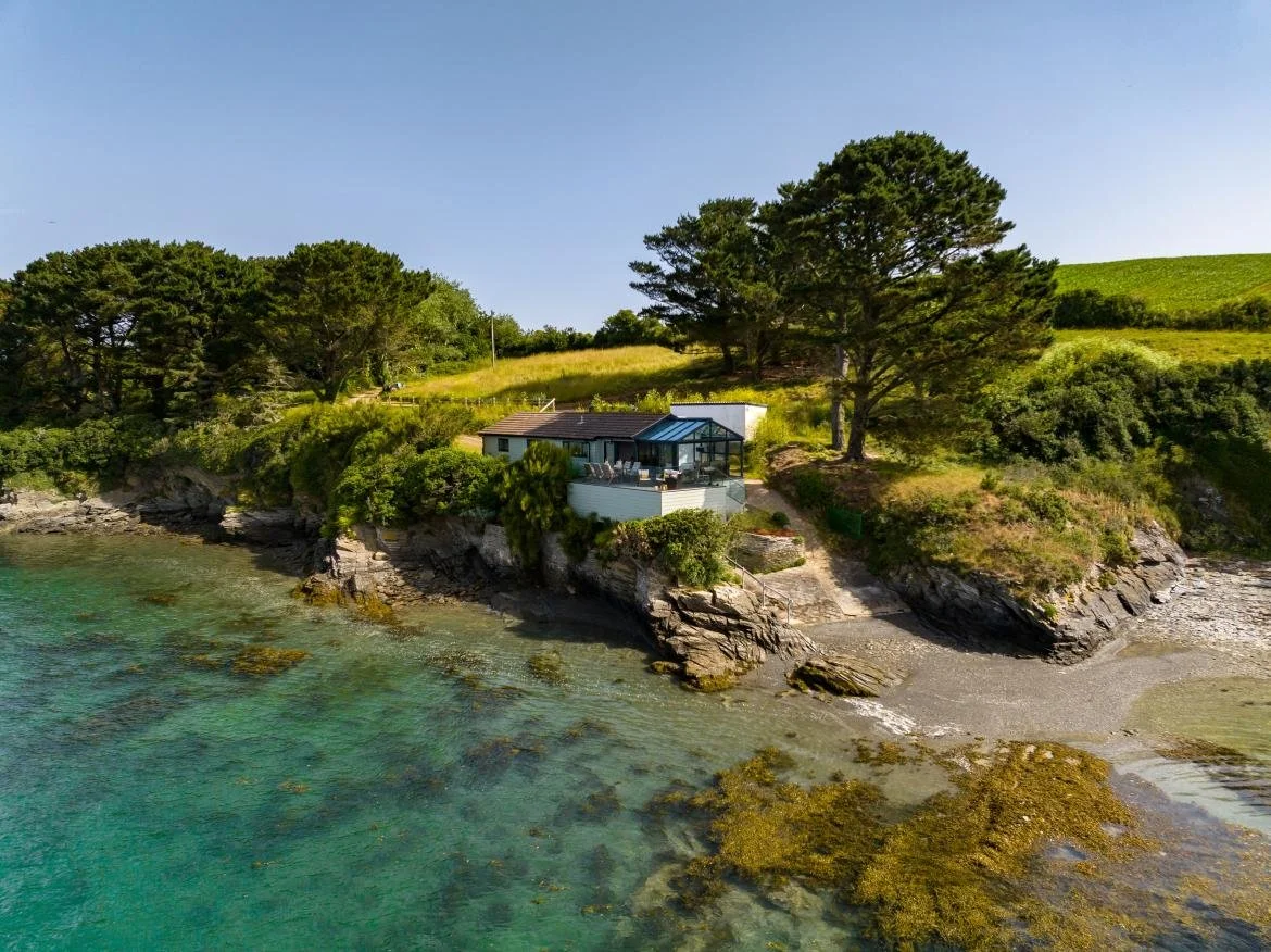 A house on a rocky shoreline with trees and green hills, under a clear blue sky.