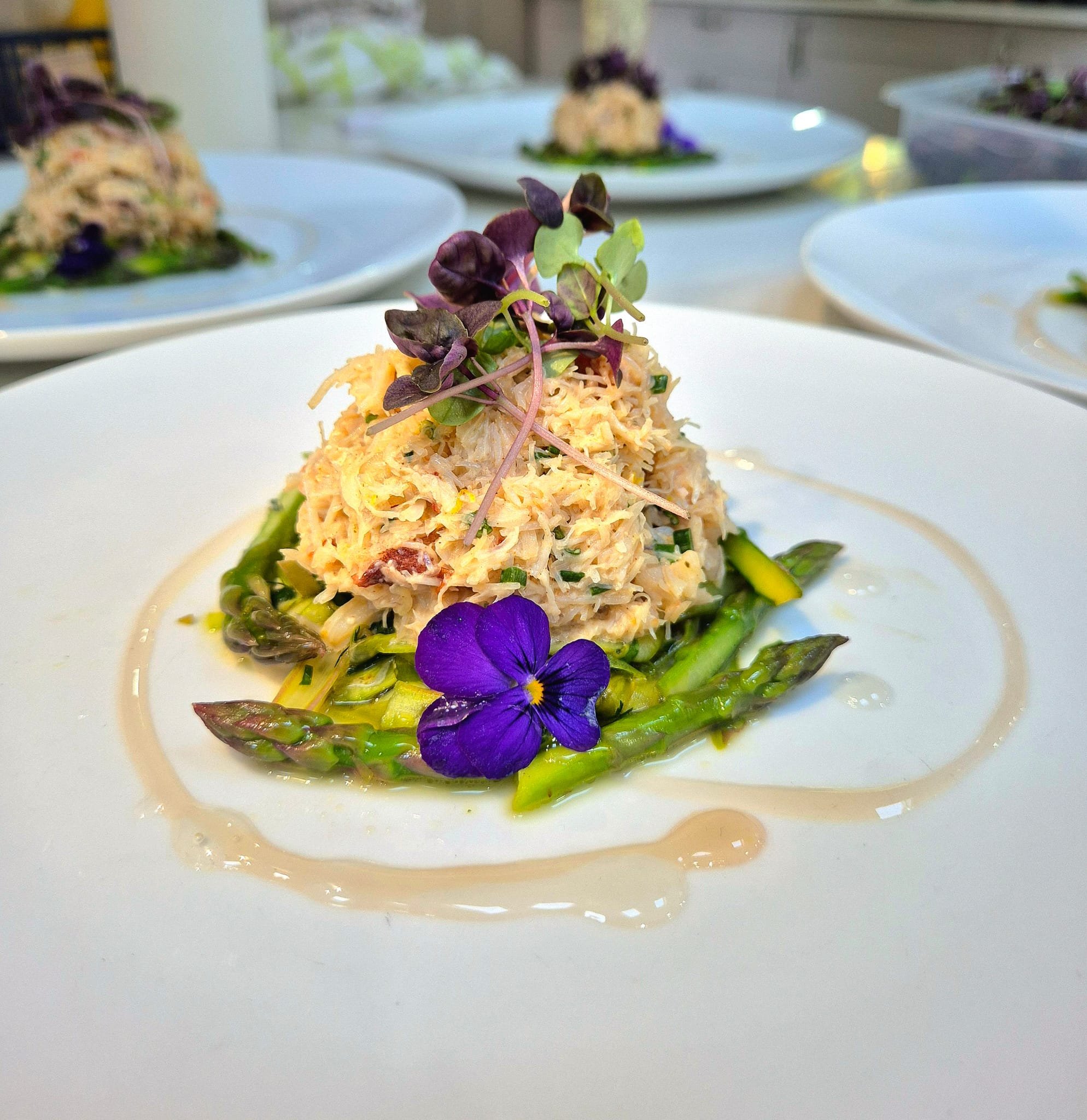 A plated dish featuring a scoop of crab salad garnished with microgreens, served over cooked asparagus with edible flowers and a drizzle of sauce.