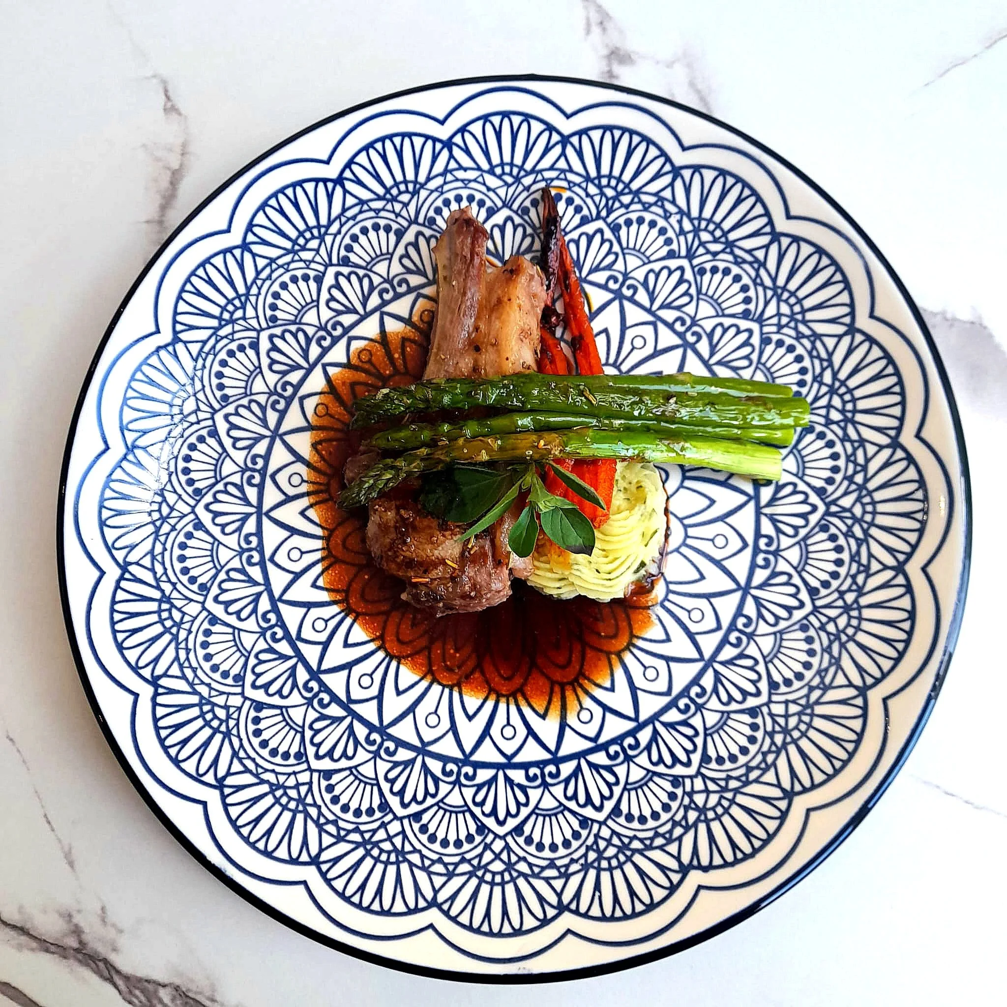 Gourmet dish with lamb chop, asparagus, mashed potatoes, carrots, and garnish on a decorative plate.