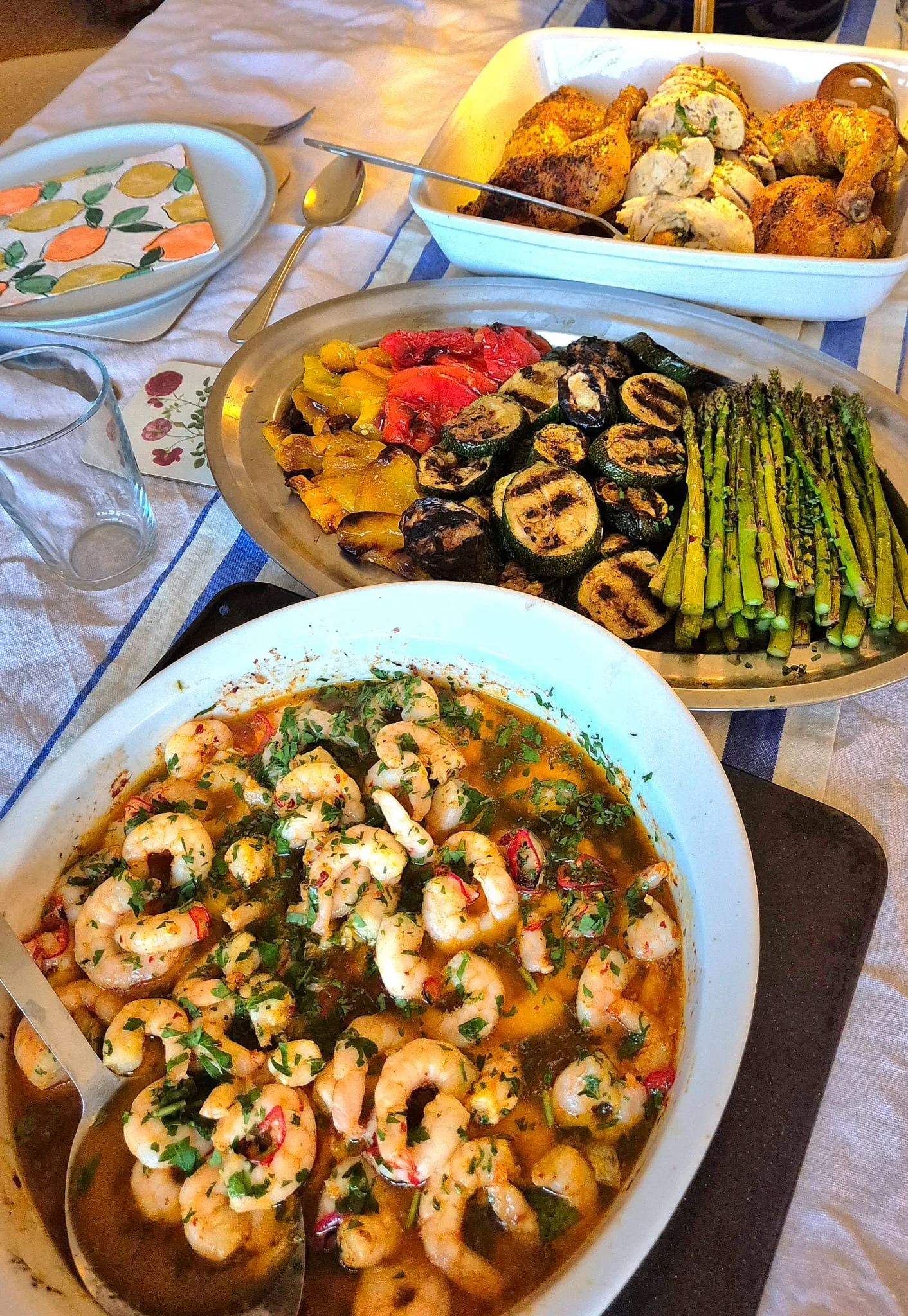 A table with various dishes including shrimp in a spicy sauce, grilled eggplant, roasted bell peppers, grilled zucchini, roasted chicken, and grilled asparagus.