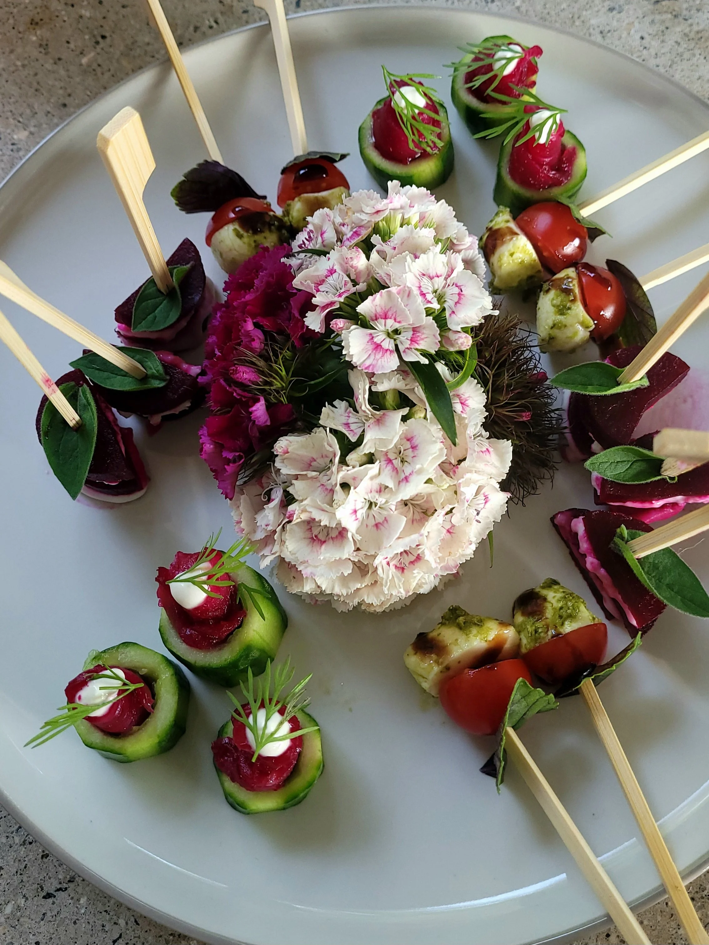 Plate with assorted appetizers, including cucumber cups with cream and dill, skewered cherry tomatoes and cheese, beet slices with herbs, and decorative pink flowers in the center.