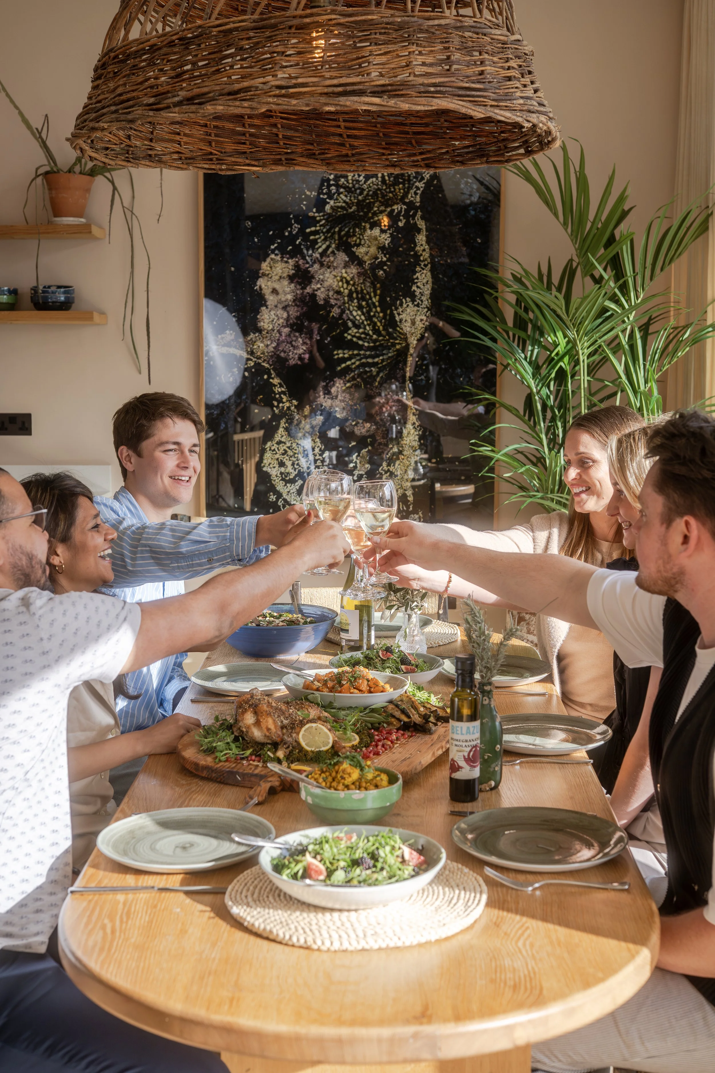 A lively group of people at a long dining table enjoying a meal together, with a variety of dishes including salads and a roasted fish, raising glasses in a toast, in a warmly lit room decorated with plants, artwork, and a large wicker lampshade.