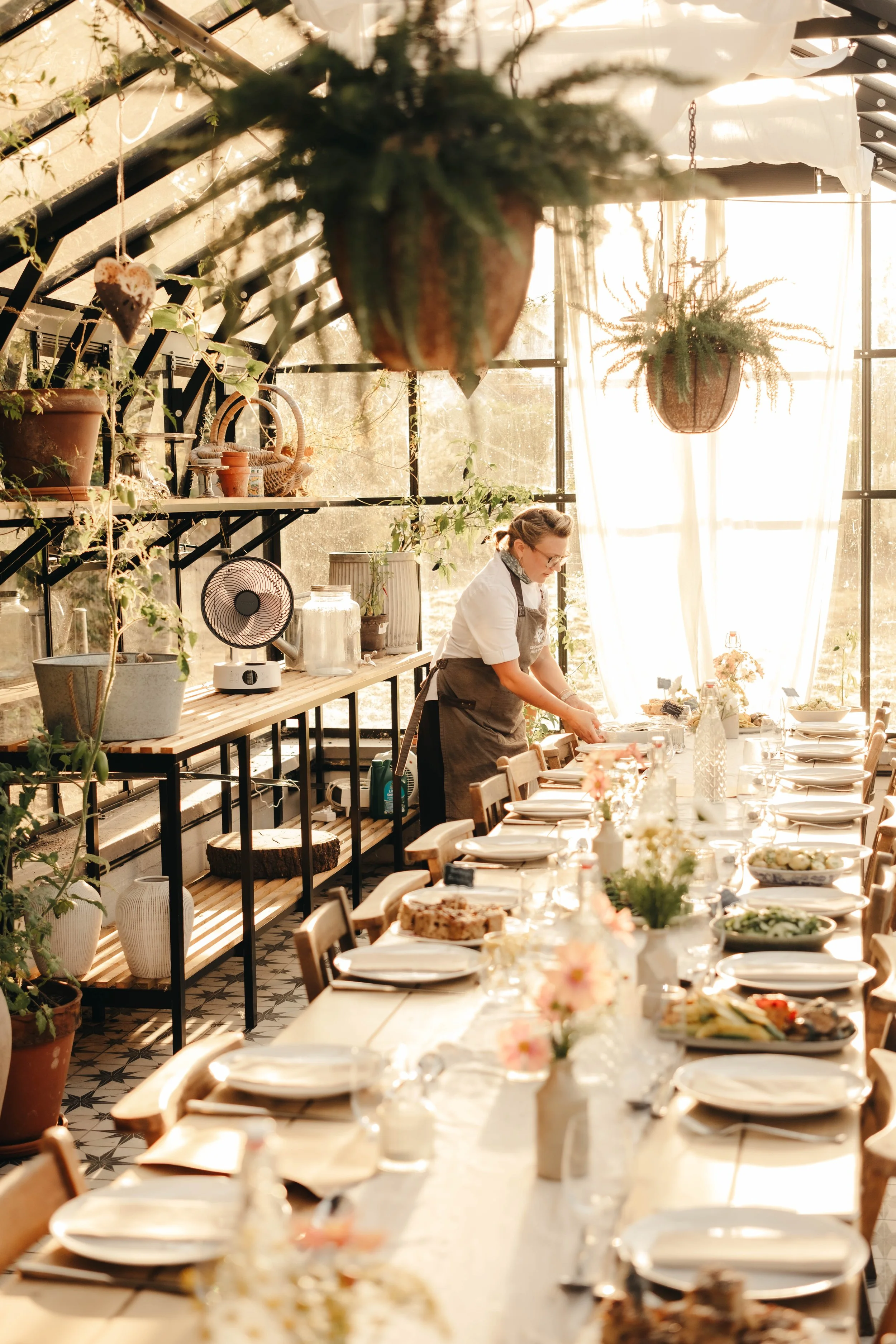 An indoor dining space with a long table set with plates, glasses, and centerpieces, a woman arranging items at the end of the table, surrounded by potted plants, hanging planters, and natural light streaming through large windows.
