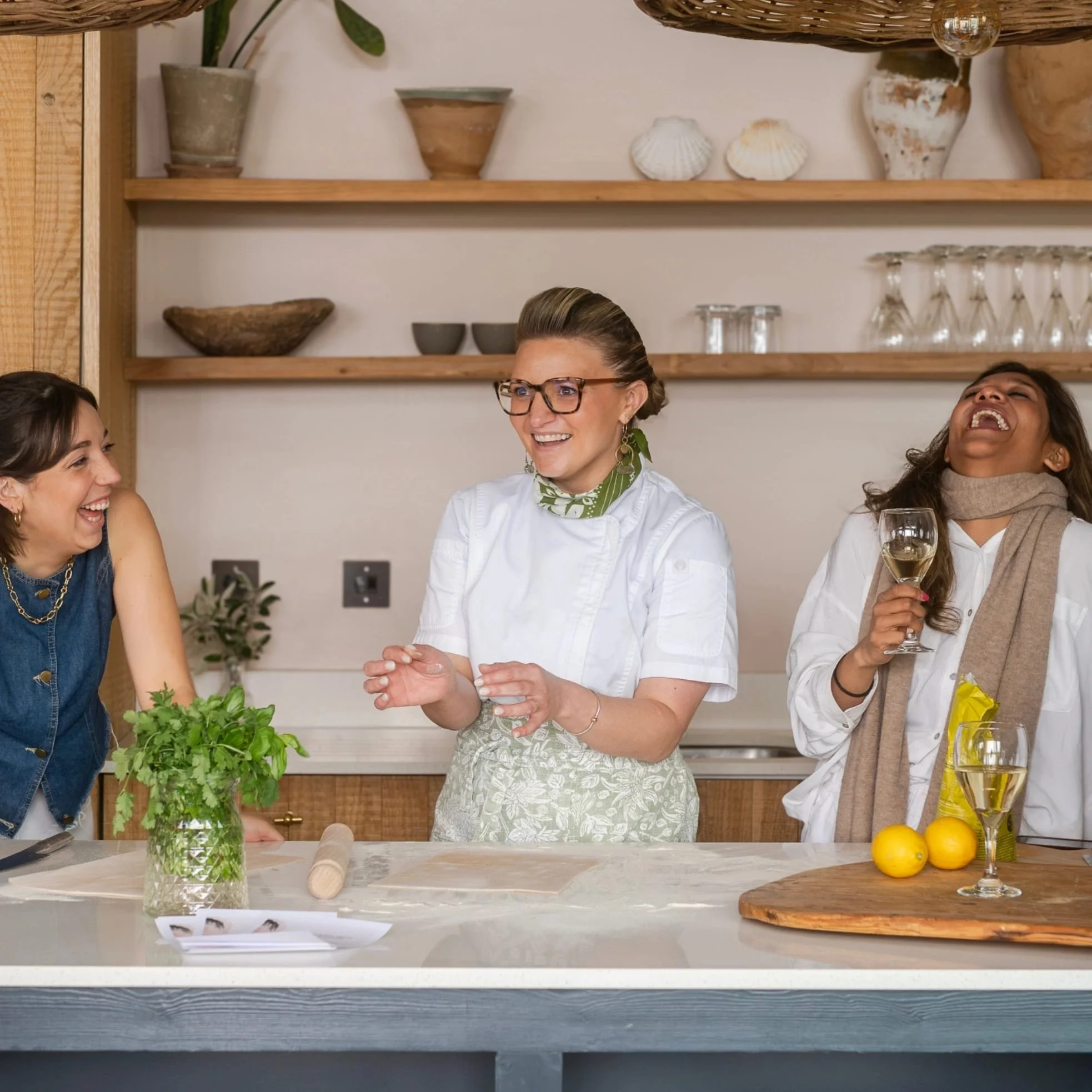 Three women laughing and enjoying a conversation in a kitchen. One woman is wearing a white chef's coat and glasses, standing behind a kitchen counter with fresh herbs and lemons. The other two women are holding wine glasses, one with white wine, and are dressed casually.