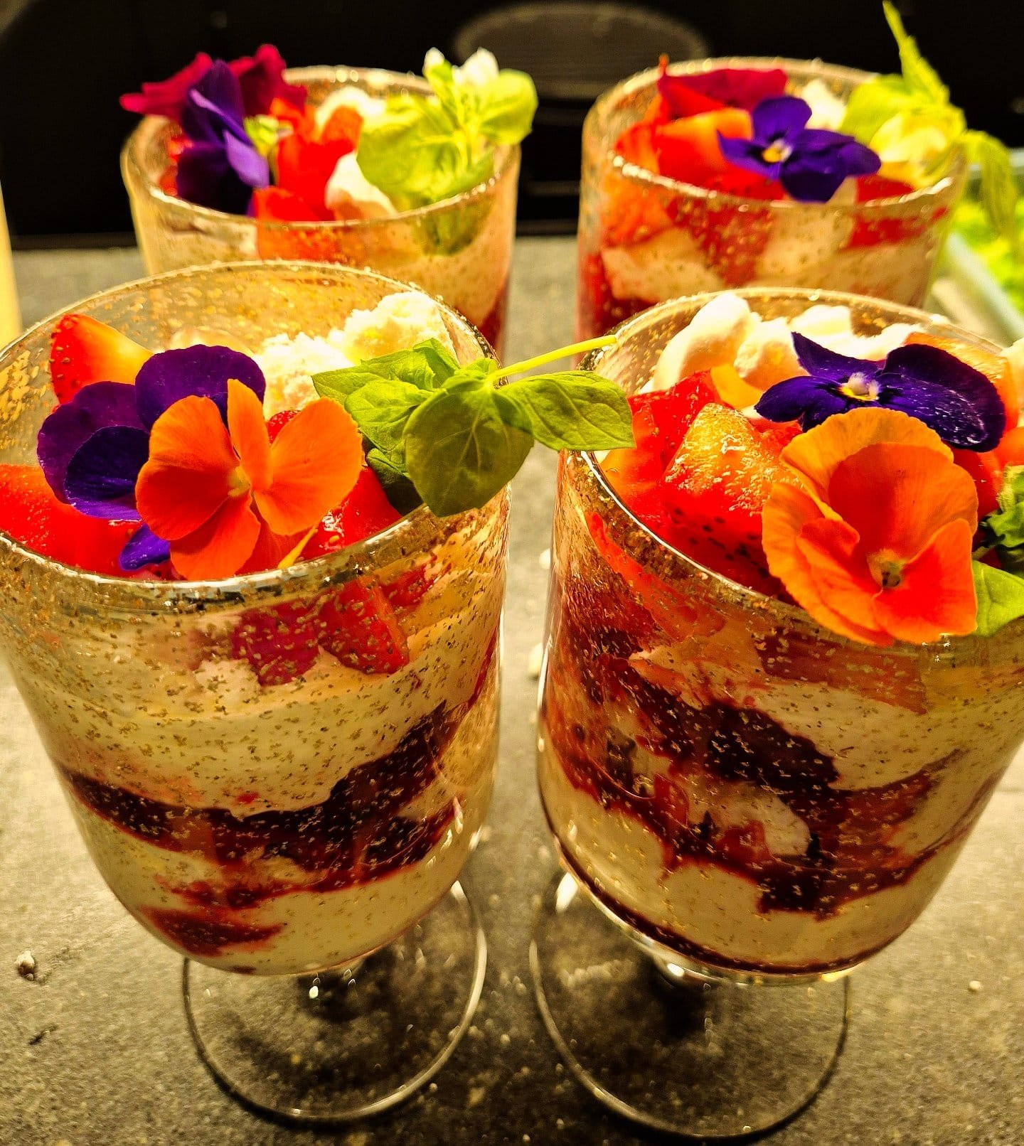 Four glasses filled with layered desserts topped with edible flowers and fresh herbs.