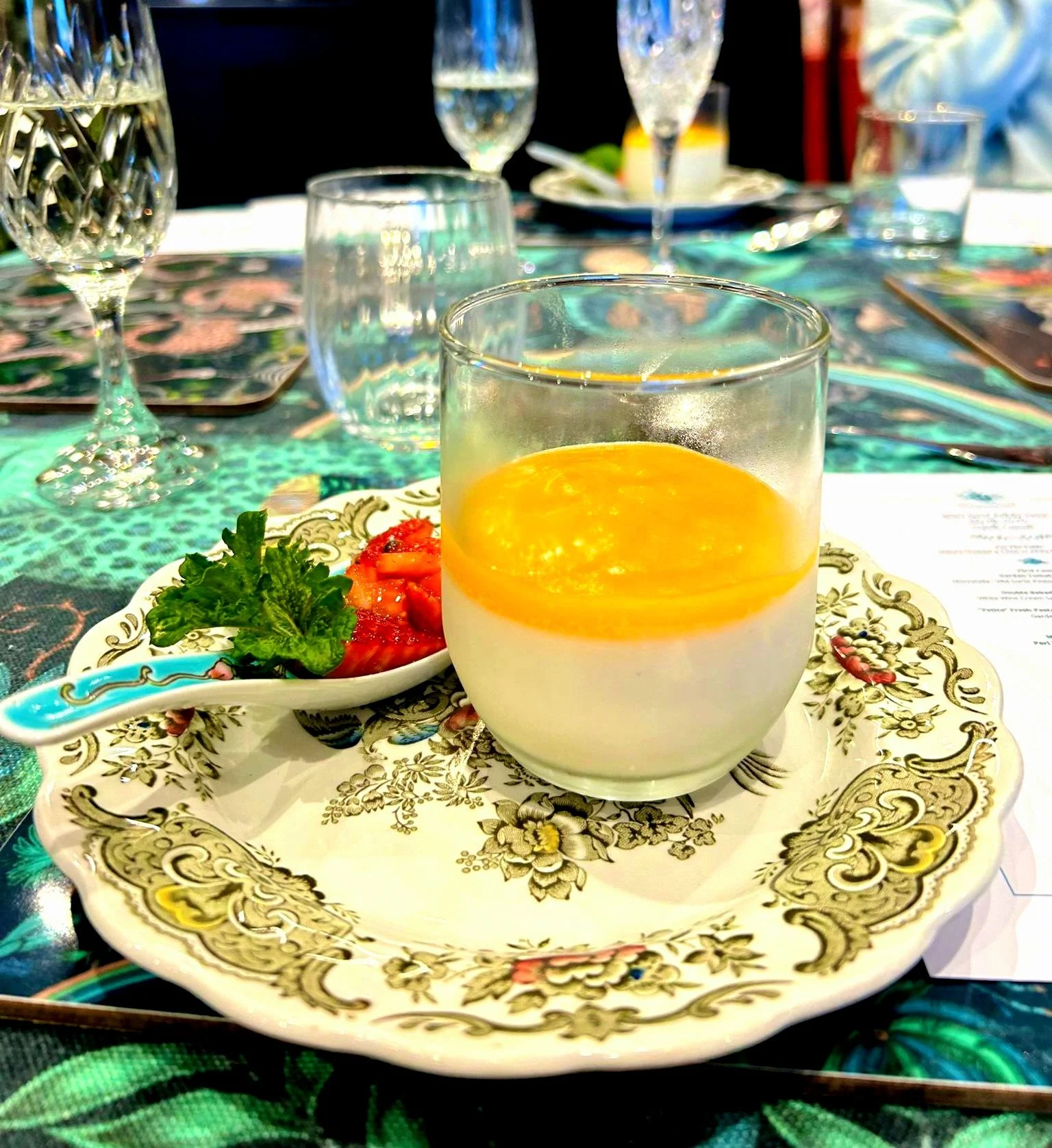 Fancy dessert plate with panna cotta topped with orange sauce, garnished with sliced strawberries and parsley, on an ornate floral plate. Background shows glasses on a patterned tablecloth.