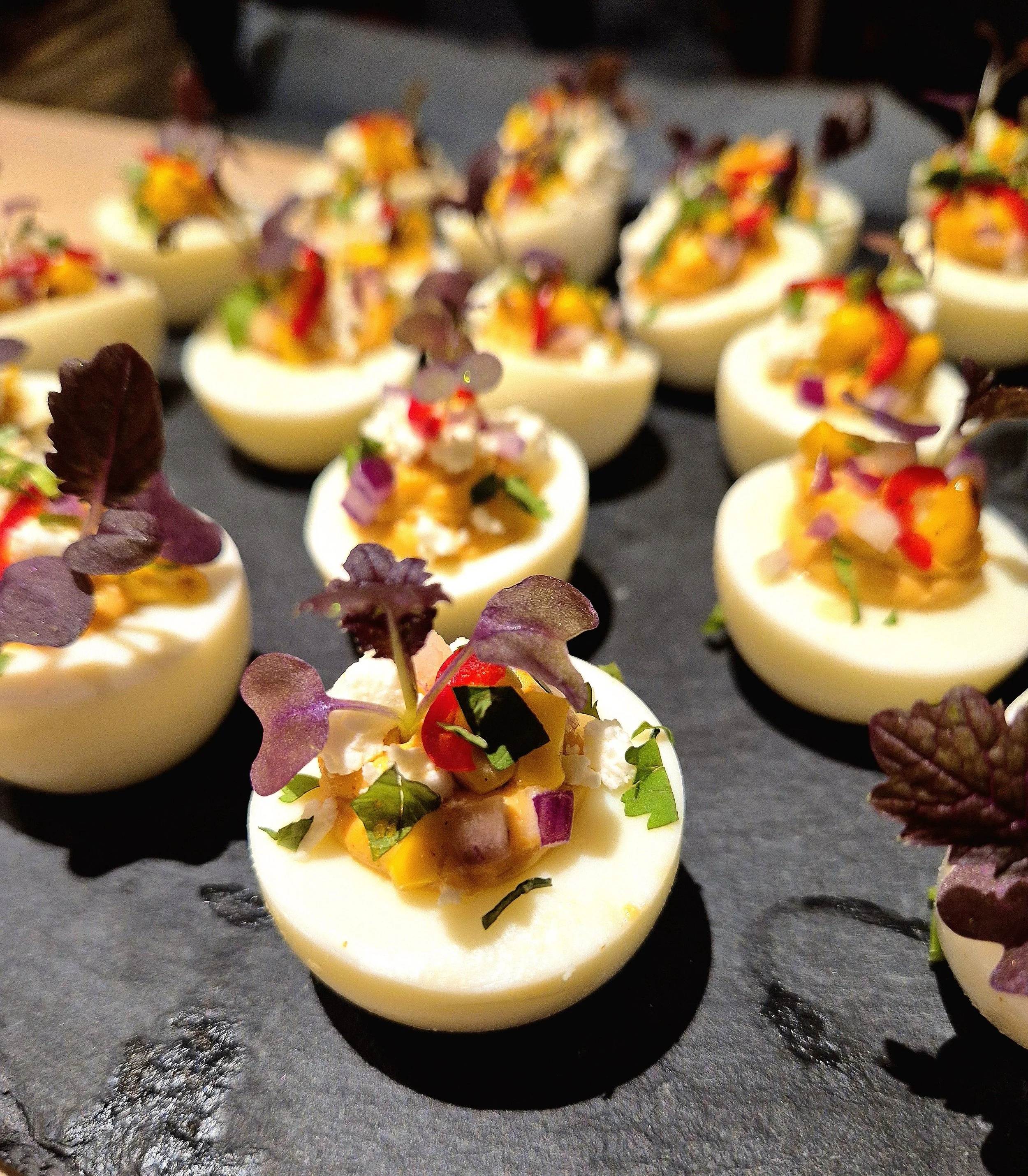 Deviled eggs topped with chopped vegetables and microgreens on a dark serving tray.