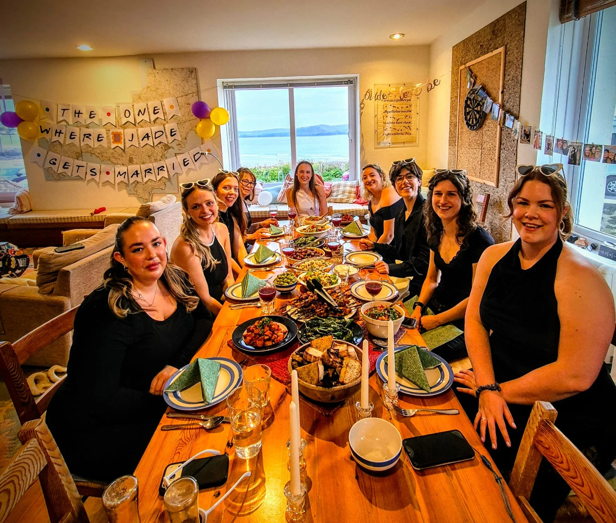 A group of women sitting around a dining table decorated for a celebration, with a banner that reads 'The One Where Jade Gets Married,' festive balloons, and a scenic view of a lake through a large window in the background.