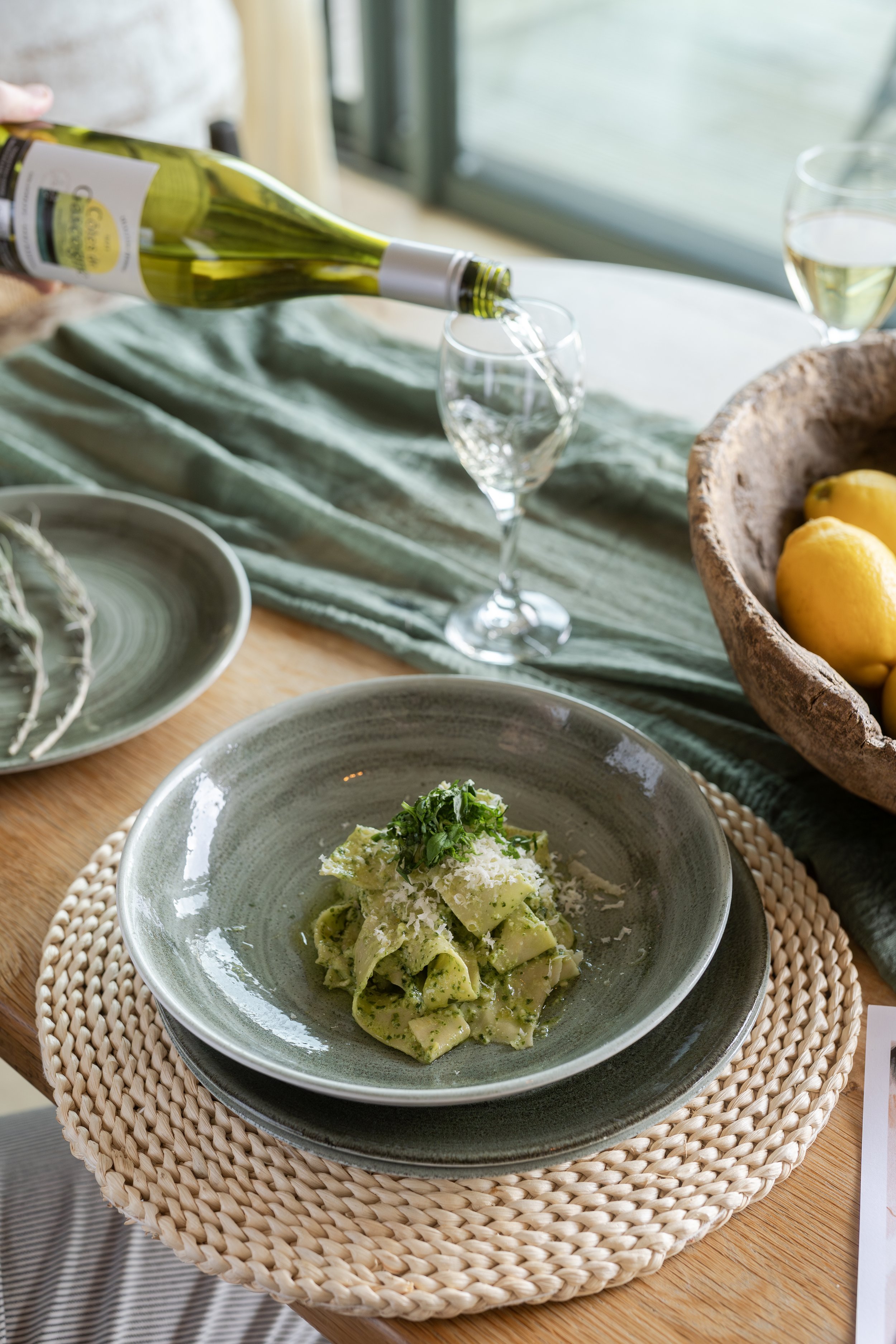 A dining table set with a plate of green pesto pasta, a hand pouring white wine into a glass, and a bowl of lemons in the background.