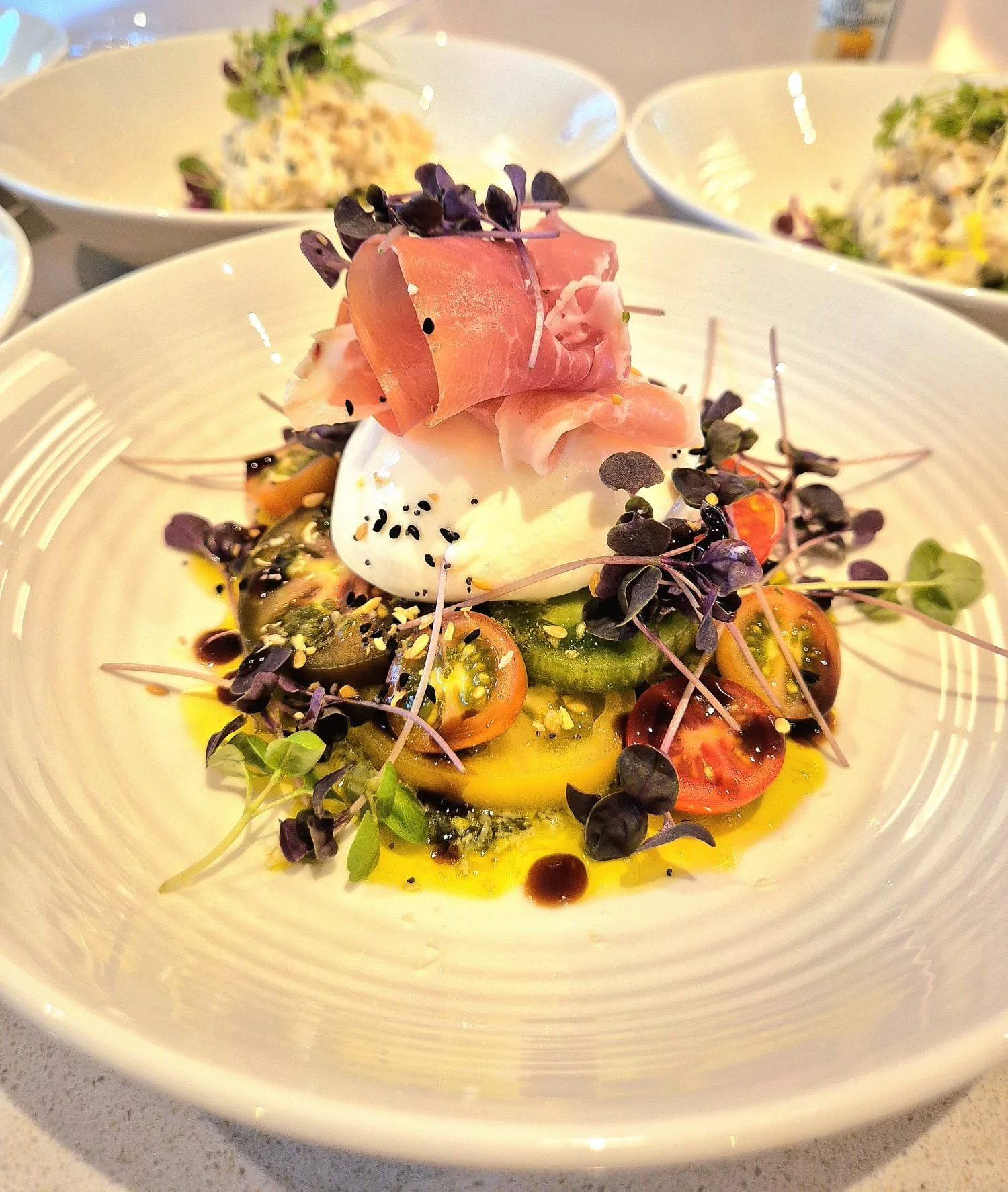 A gourmet salad with cherry tomatoes, cucumber slices, fresh mozzarella, microgreens, black sesame seeds, and balsamic glaze, topped with prosciutto and a poached egg.