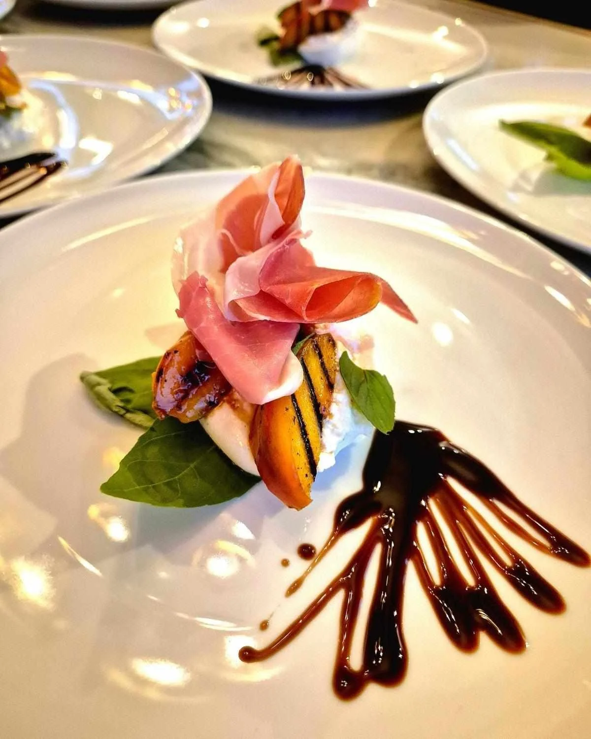 Elegant dish featuring prosciutto, grilled peach slices, fresh mozzarella, basil leaves, and balsamic glaze on a white plate.