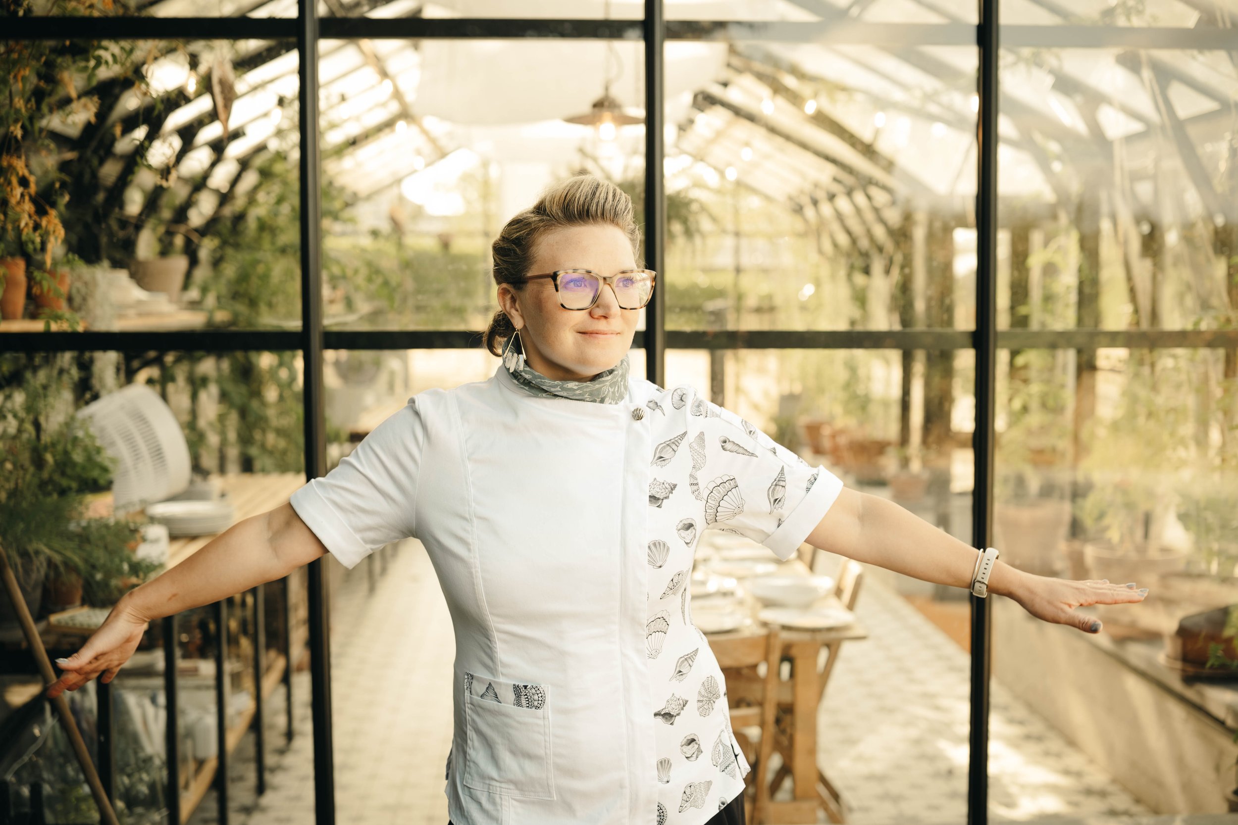 A woman in a white shell-patterned uniform and glasses stands with her arms extended in a greenhouse filled with plants and gardening supplies.