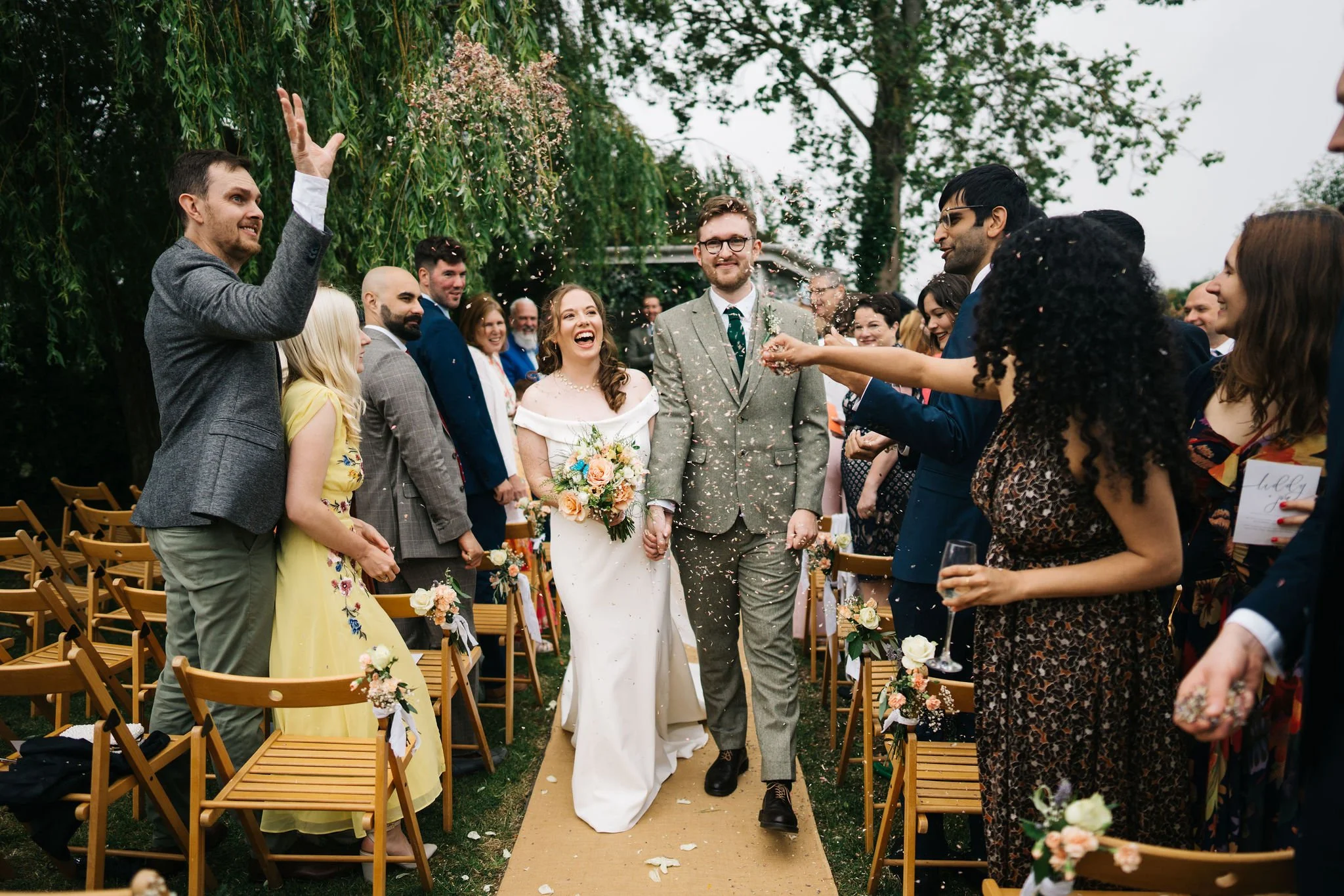 A bride and groom walk down the aisle at their outdoor wedding, surrounded by guests, with friends throwing confetti in the air.