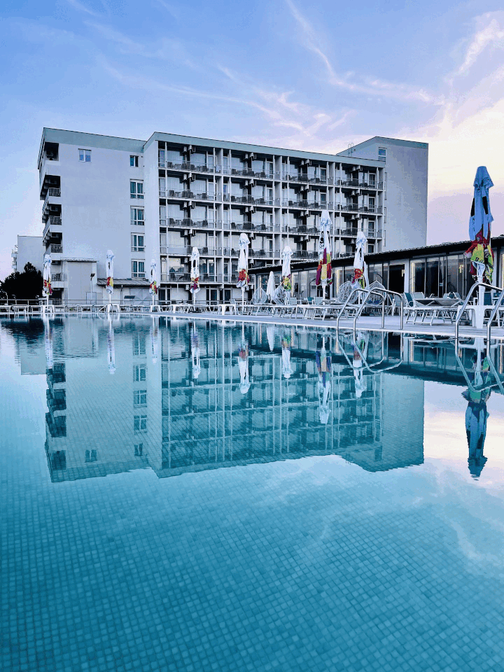 An outdoor swimming pool with lounge chairs and closed umbrellas in front of a hotel building during the evening, with the building's reflection visible in the pool.