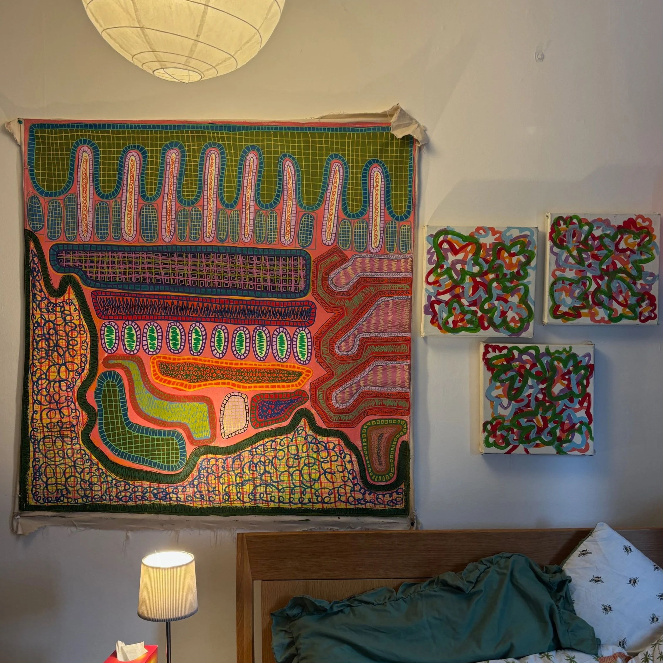 Colorful abstract artwork hanging on a white wall above a wooden bed with blue bedding and pillows, illuminated by a bedside lamp, with a paper tissue box on a small table.