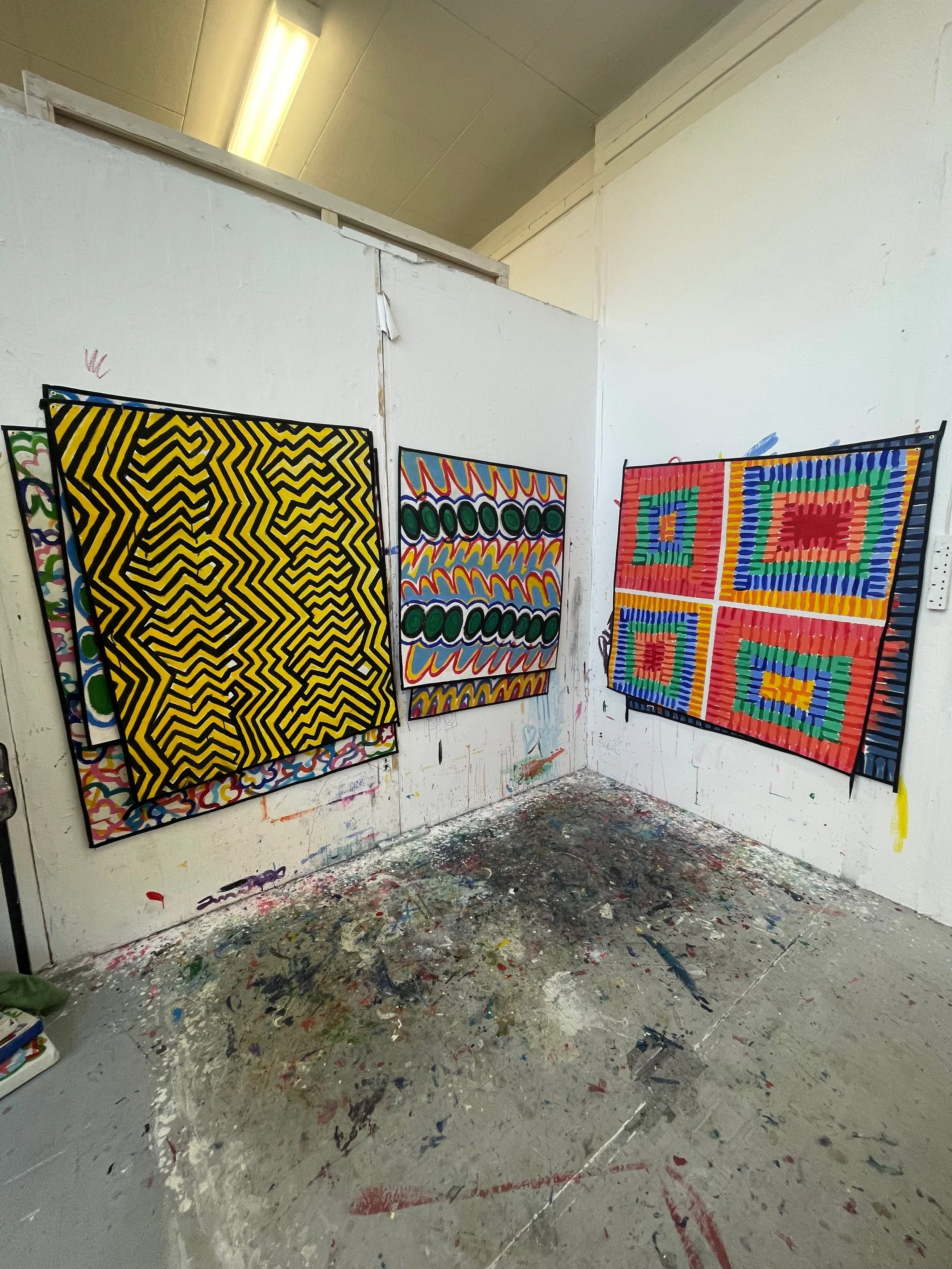 Three colorful geometric abstract paintings hanging on a white wall in an art studio, with a paint-splattered floor.