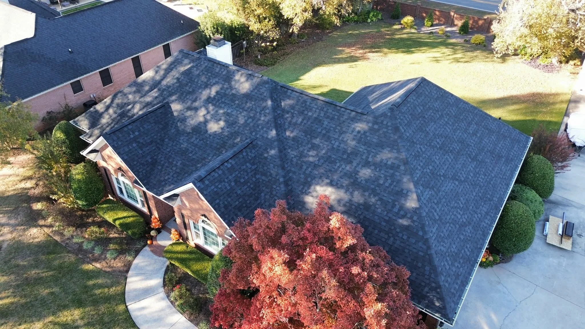 Aerial view of a suburban house in Warner Robins Georgia with a dark shingle roof, brick exterior, and front yard with a curved walkway, surrounded by trees and shrubs, including a prominent red-leafed tree in the front yard.