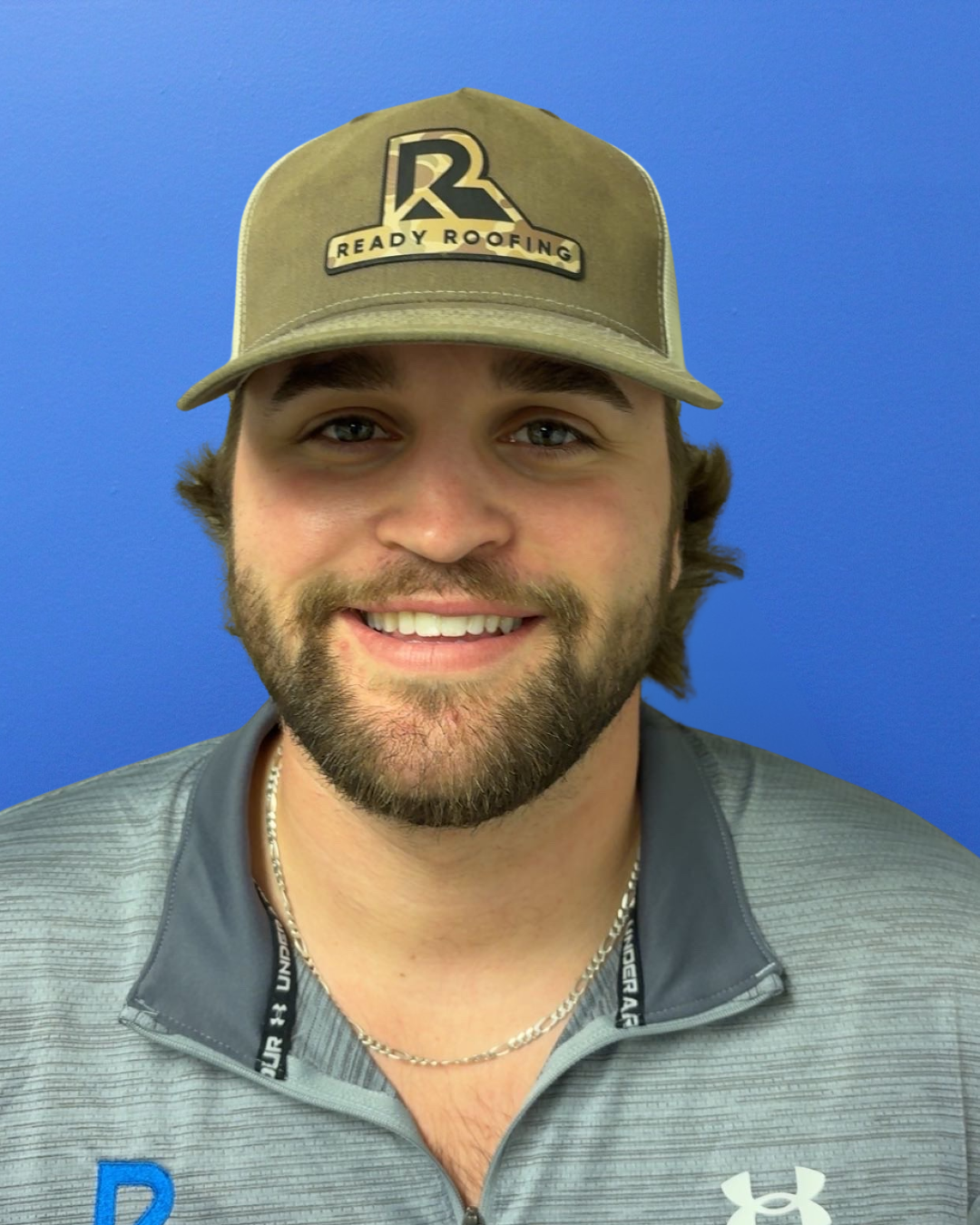 A smiling man wearing a beige cap with the logo 'Ready Roofing' and a gray Under Armour jacket, standing against a blue background.