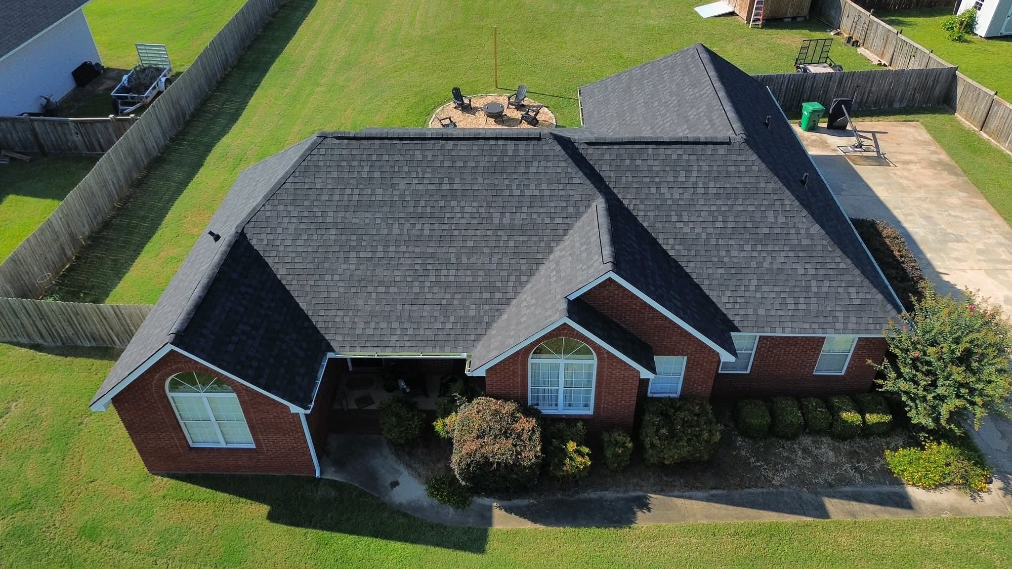Aerial view of a brick house in Kathleen Georgia with a dark shingle roof, surrounded by a lawn and fenced backyard, featuring a patio with outdoor furniture, a small garden, and a paved driveway with a green trash bin.