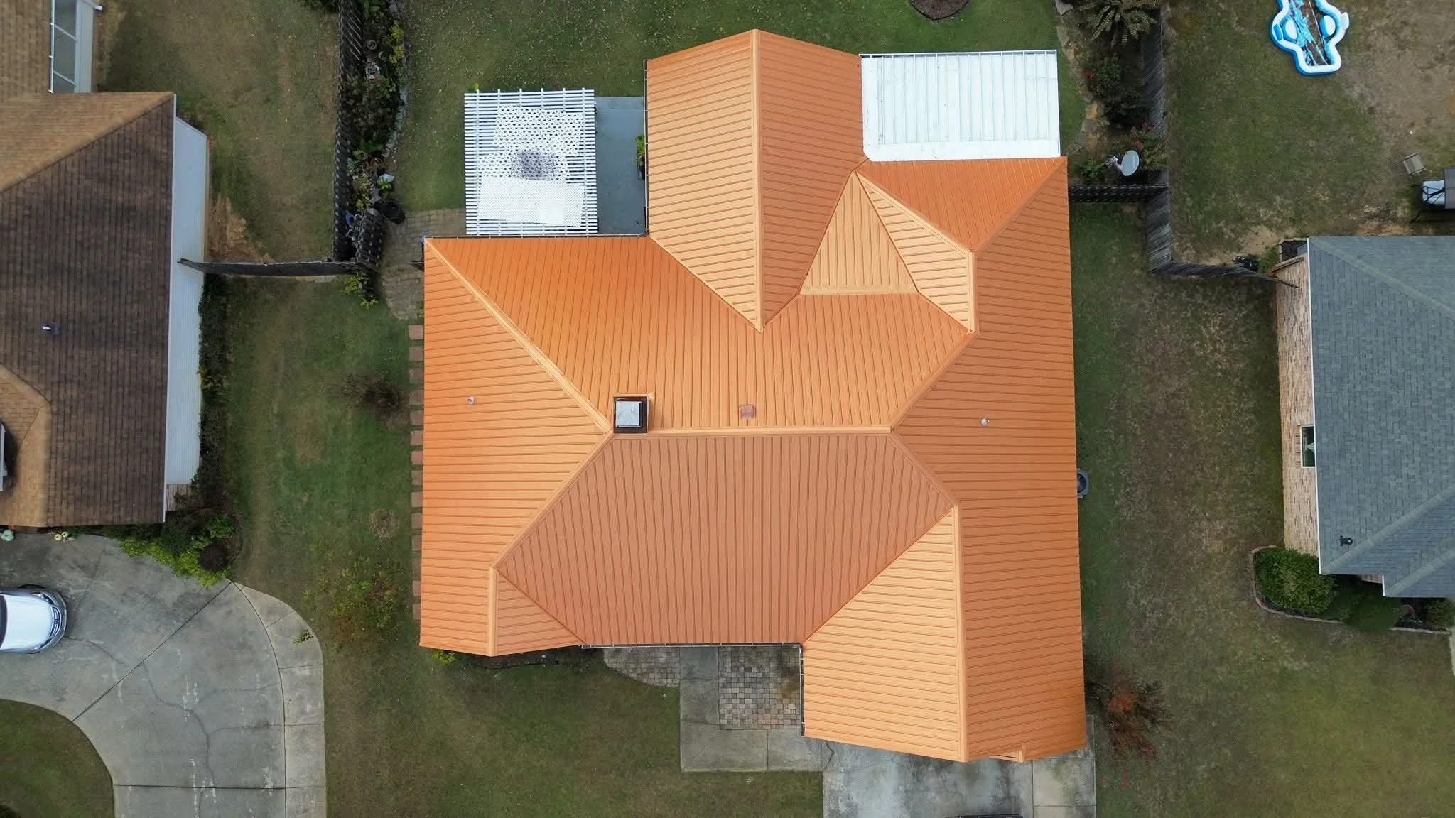 Aerial view of a house with a reddish-orange metal roof, multiple peaks and valleys. The house is surrounded by a lawn and neighboring houses. There is a small concrete driveway with a white vehicle parked on it, and a paved pathway