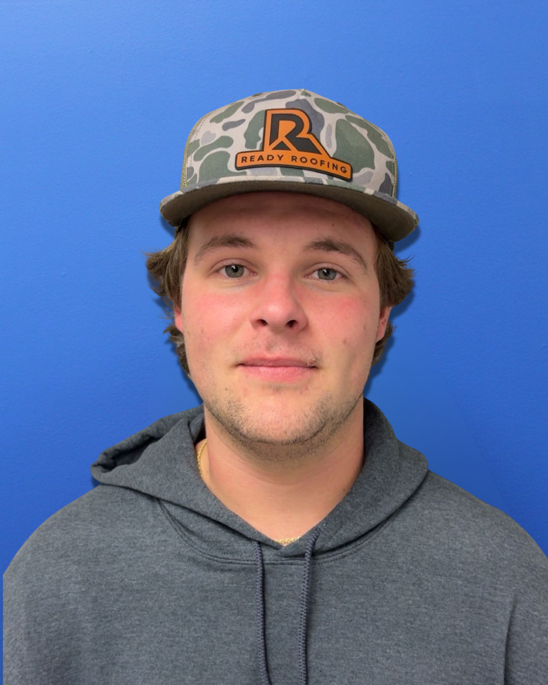A young man with light skin, brown hair, wearing a gray hoodie, and a camouflage baseball cap with an orange and black logo that reads 'Ready Roofing,' standing in front of a plain blue wall.