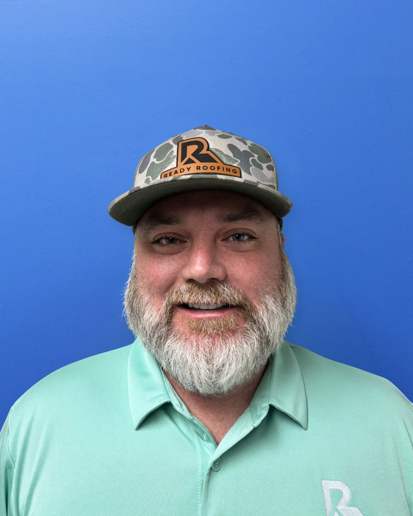 A smiling man with a gray beard wearing a camouflage cap with an orange and black logo that says 'Ready Roofing' and a light green collared shirt against a blue background.