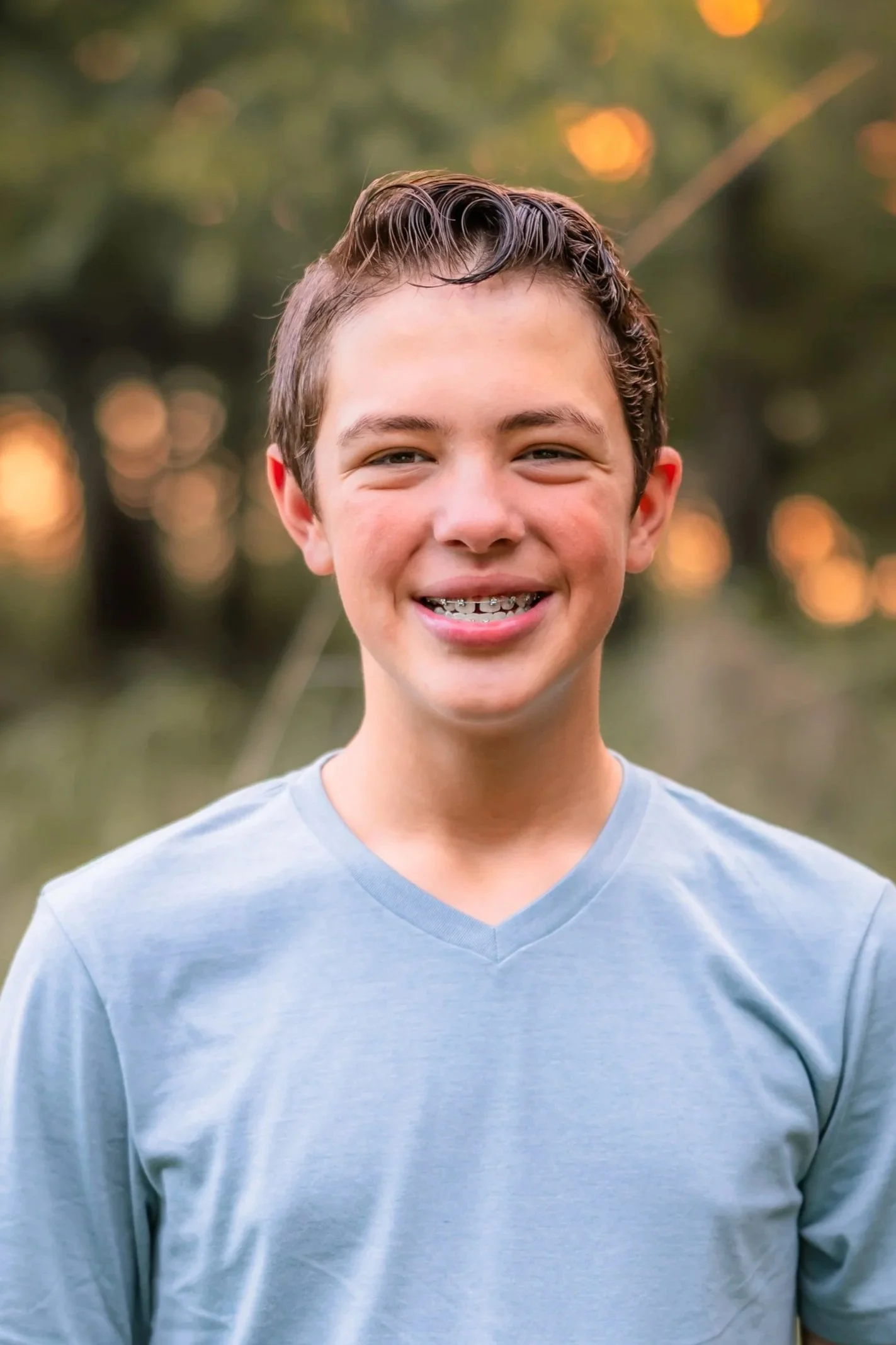 Close-up of a smiling teenage boy with braces, wearing a light blue V-neck T-shirt, outdoors with blurred trees and warm sunset in background.