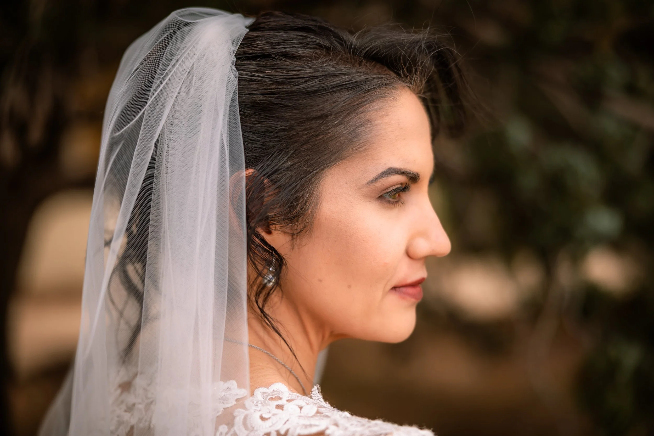 A close-up profile of a woman with dark wavy hair wearing a white veil with lace detail, outdoors with a blurred natural background.