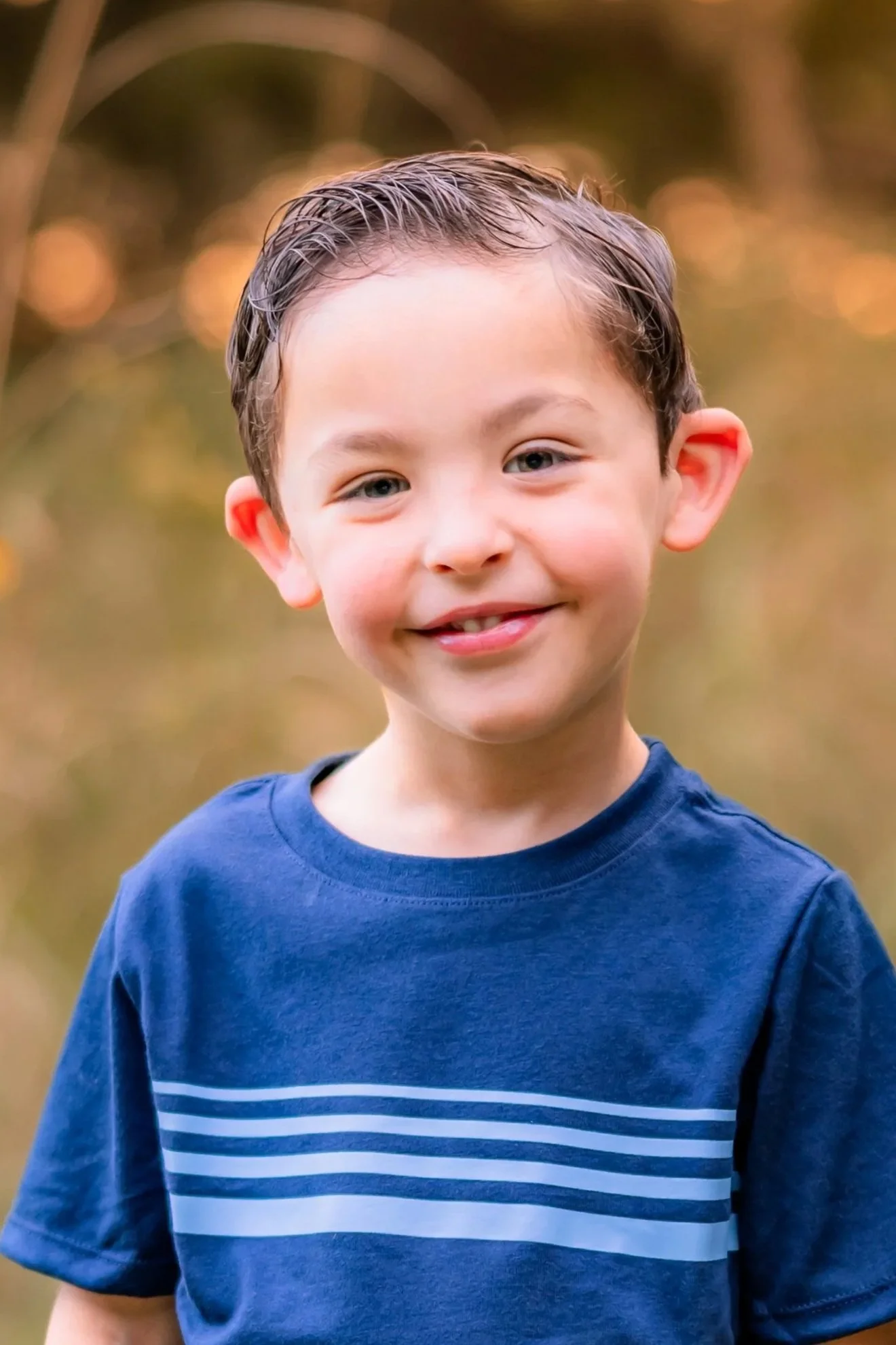 Close-up of a smiling young boy with wet hair wearing a dark blue T-shirt with light blue horizontal stripes, outdoors during fall. Blurred fall foliage background.