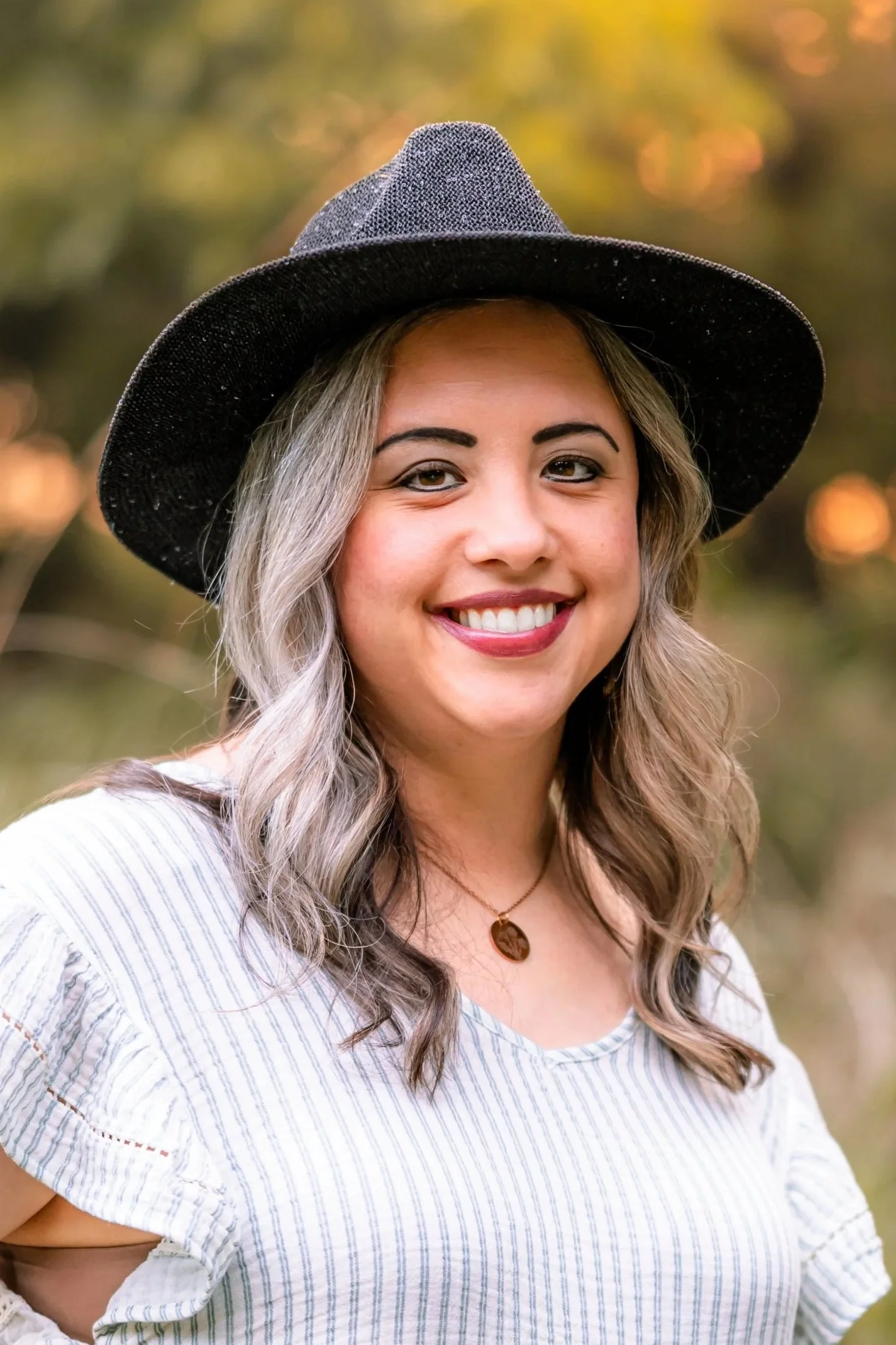 A woman with long, light gray hair wearing a black wide-brimmed hat and a white striped top, smiling outdoors with blurred warm-toned background.