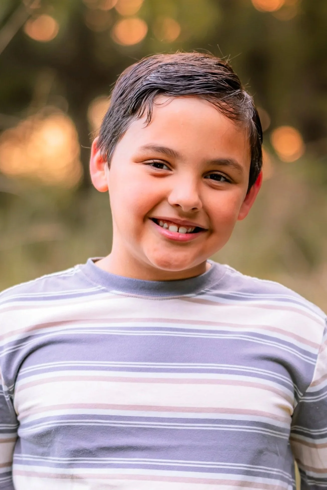 A young boy with short, wet hair smiling outdoors at sunset or sunrise, wearing a striped gray and white T-shirt.
