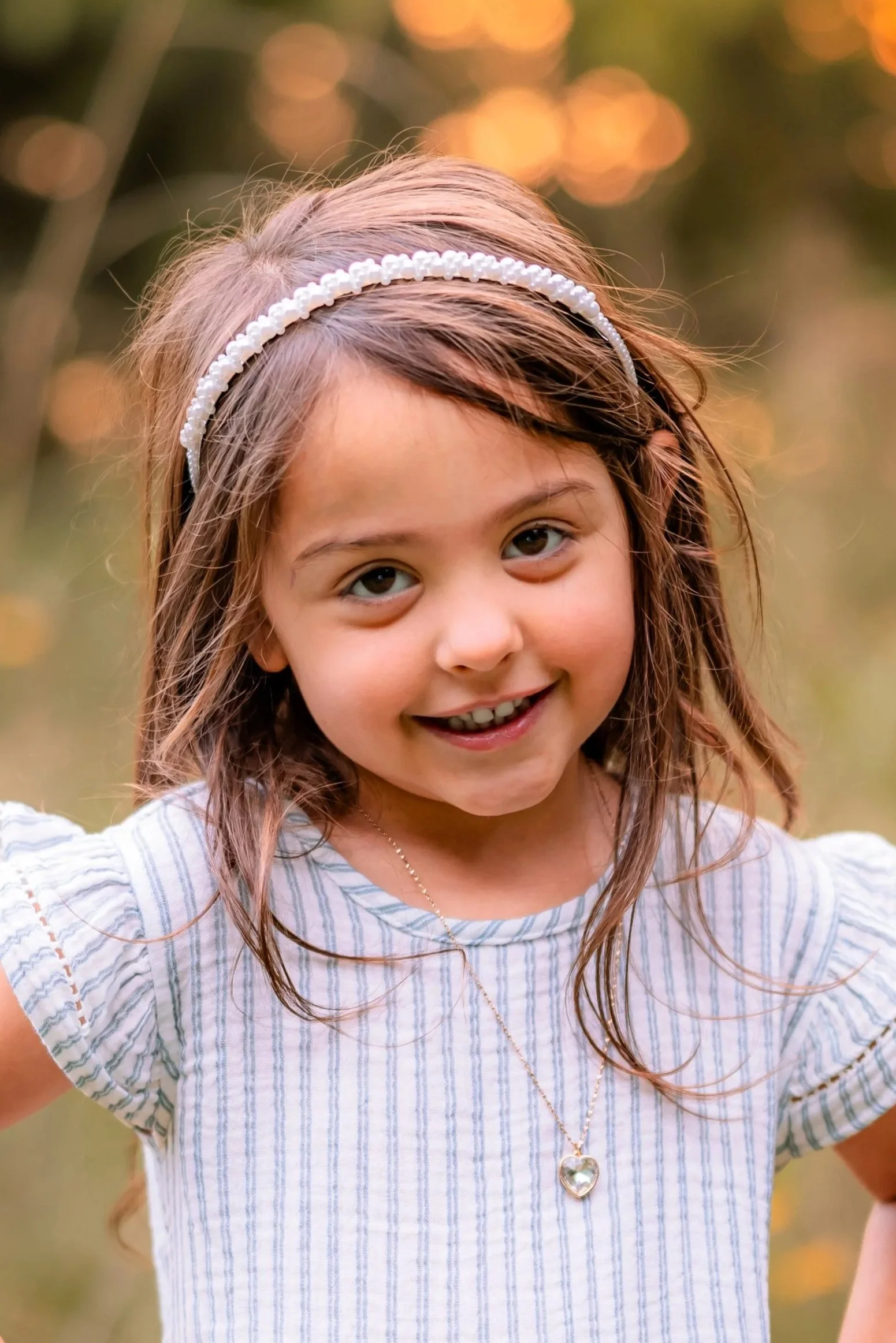 A young girl with long brown hair and a pearl headband smiling outdoors during sunset.