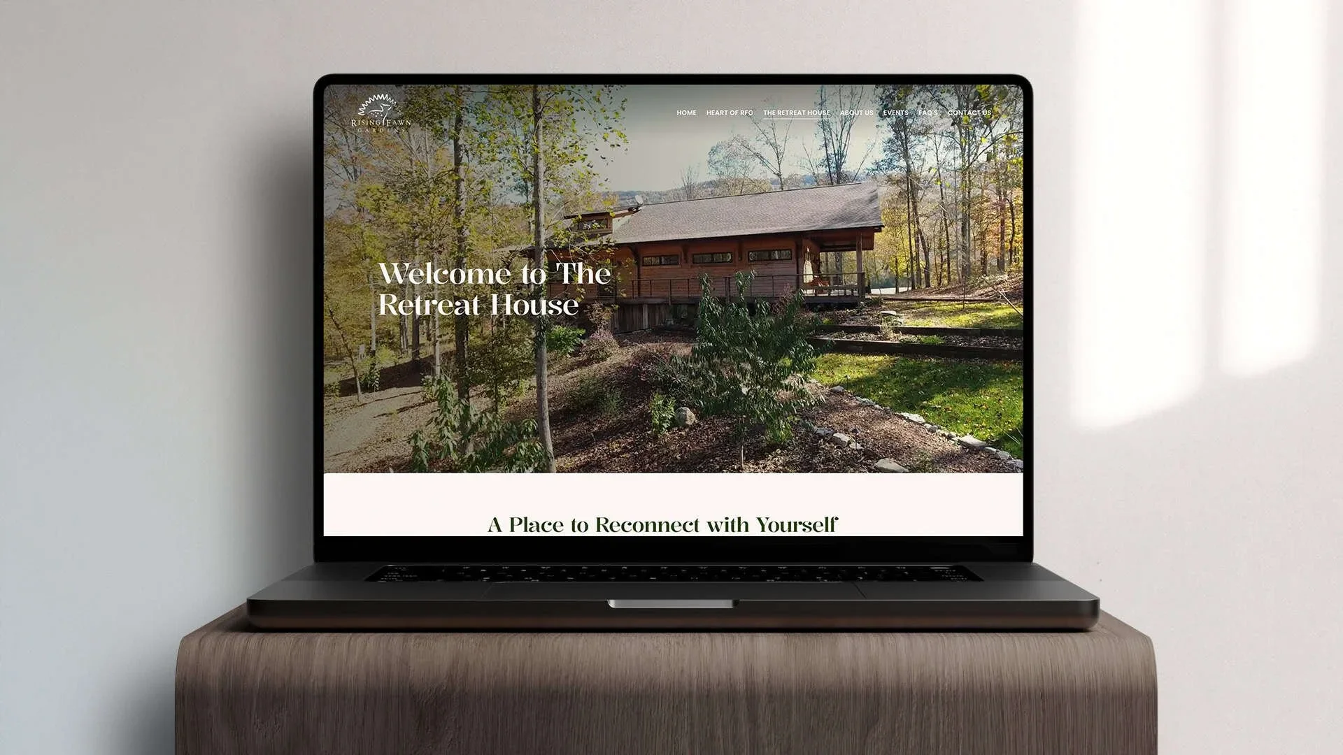 A laptop on a wooden table displaying a website for a retreat house, with an image of a wooden house in a wooded area and the text "Welcome to The Retreat House" and "A Place to Reconnect with Yourself".