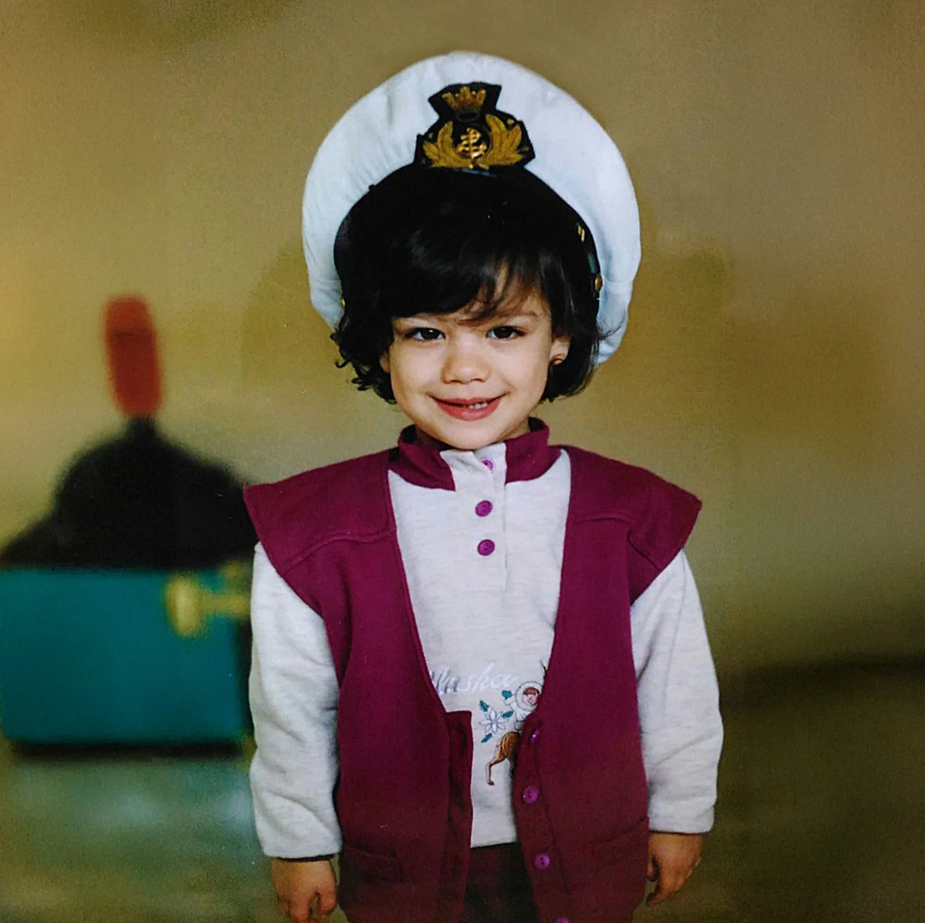 Rosie Di Lecce as a child wearing a white captain style hat and burgundy vest indoors.