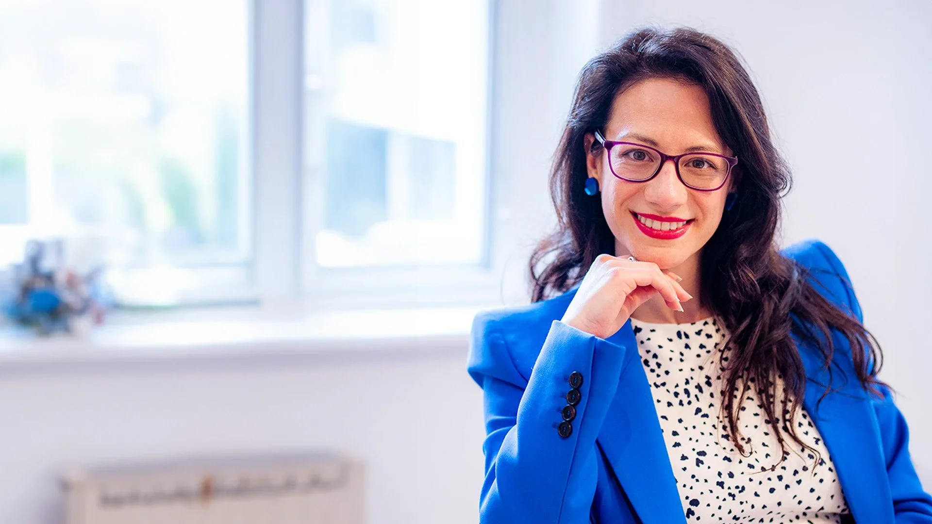 Rosie Di Lecce smiling by a window in a blue blazer and patterned dress.